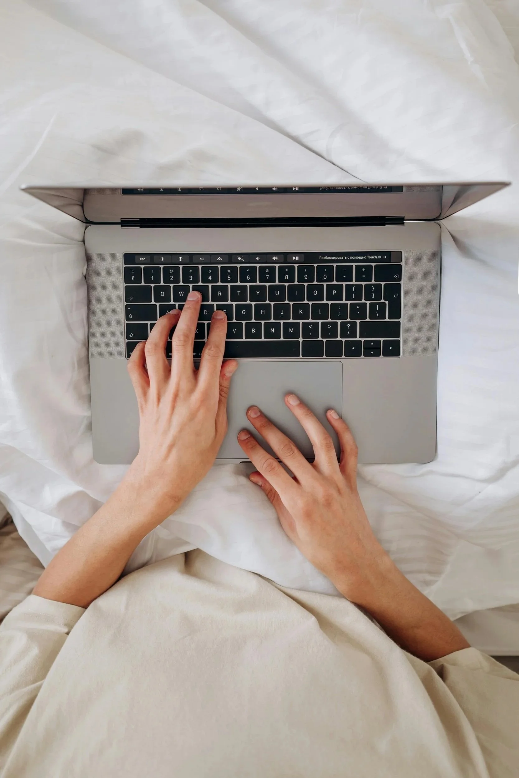 A person is typing on a MacBook laptop placed on a bed, with their hands visible.