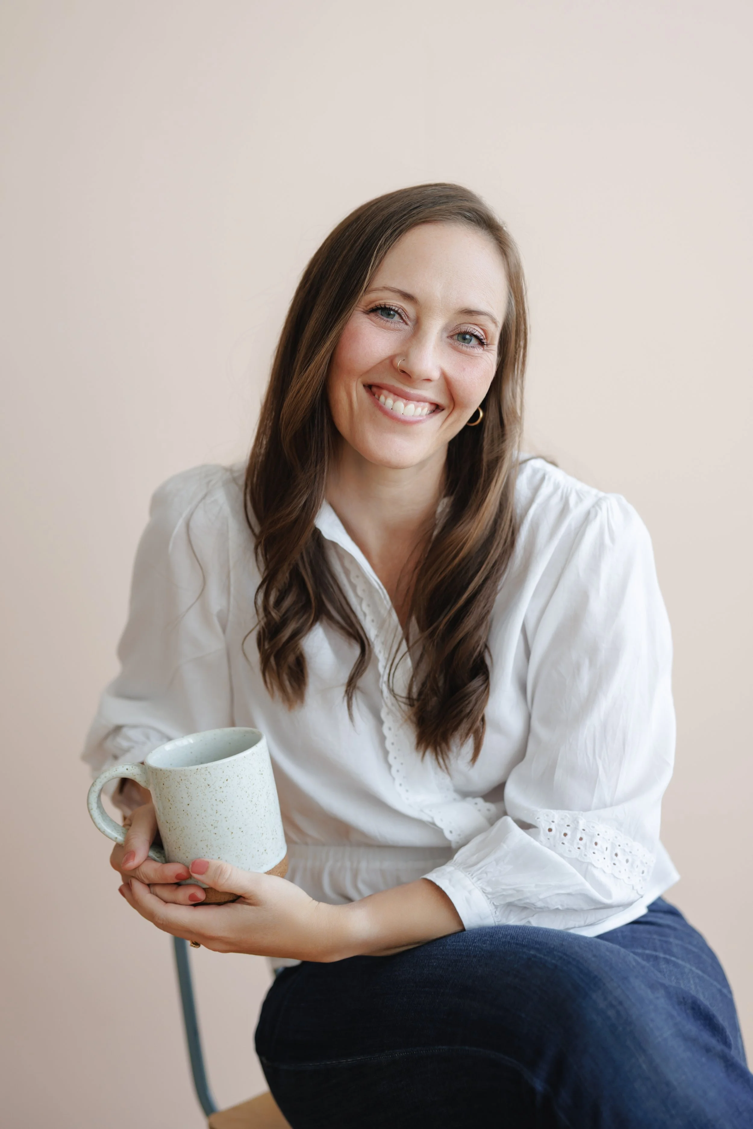 A woman with long brown hair, wearing a white blouse and dark jeans, smiling and holding a mug while sitting on a chair against a neutral background.