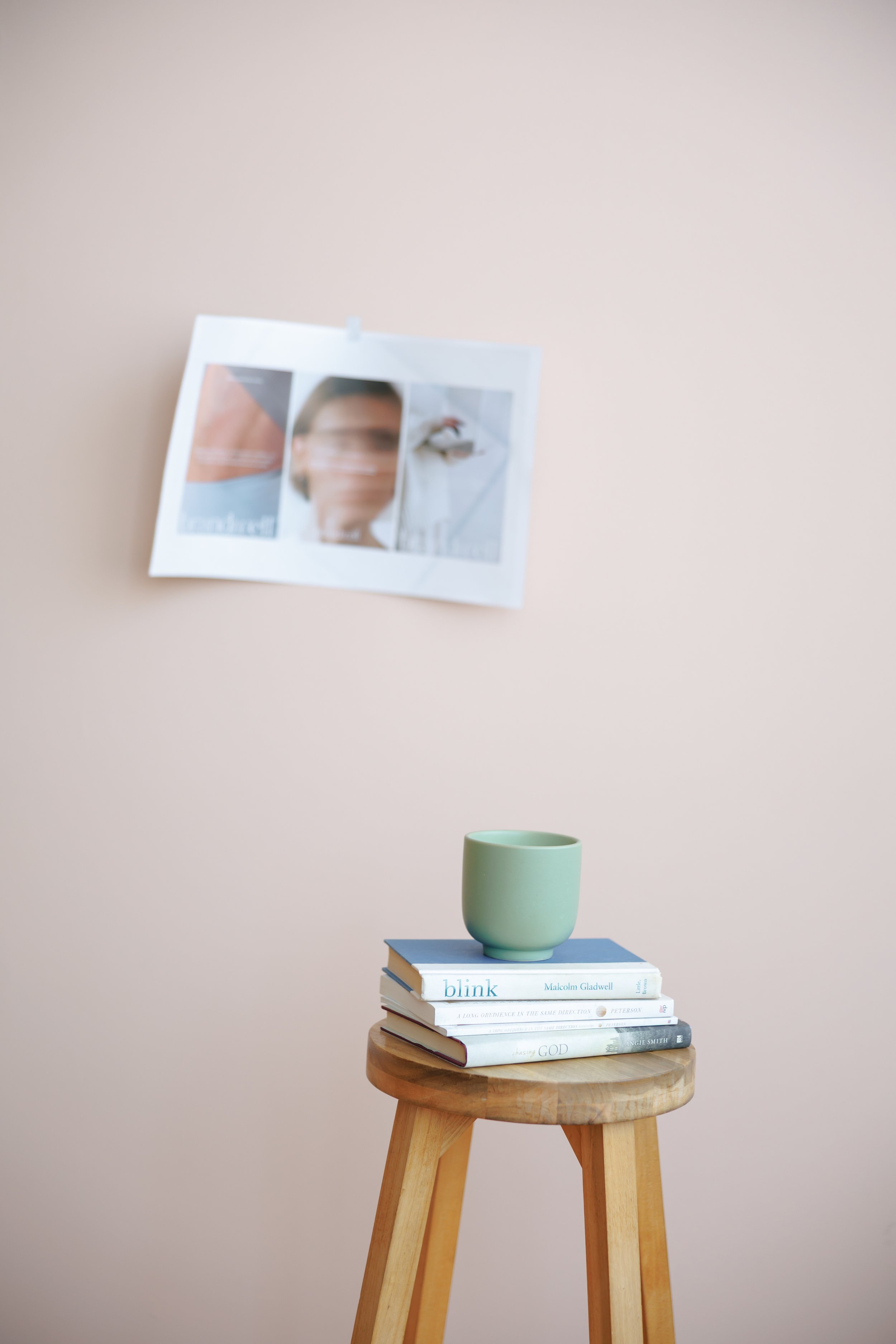 A wooden stool with a stack of books and a green mug on top, against a pale pink wall with a blurred poster hanging on it.