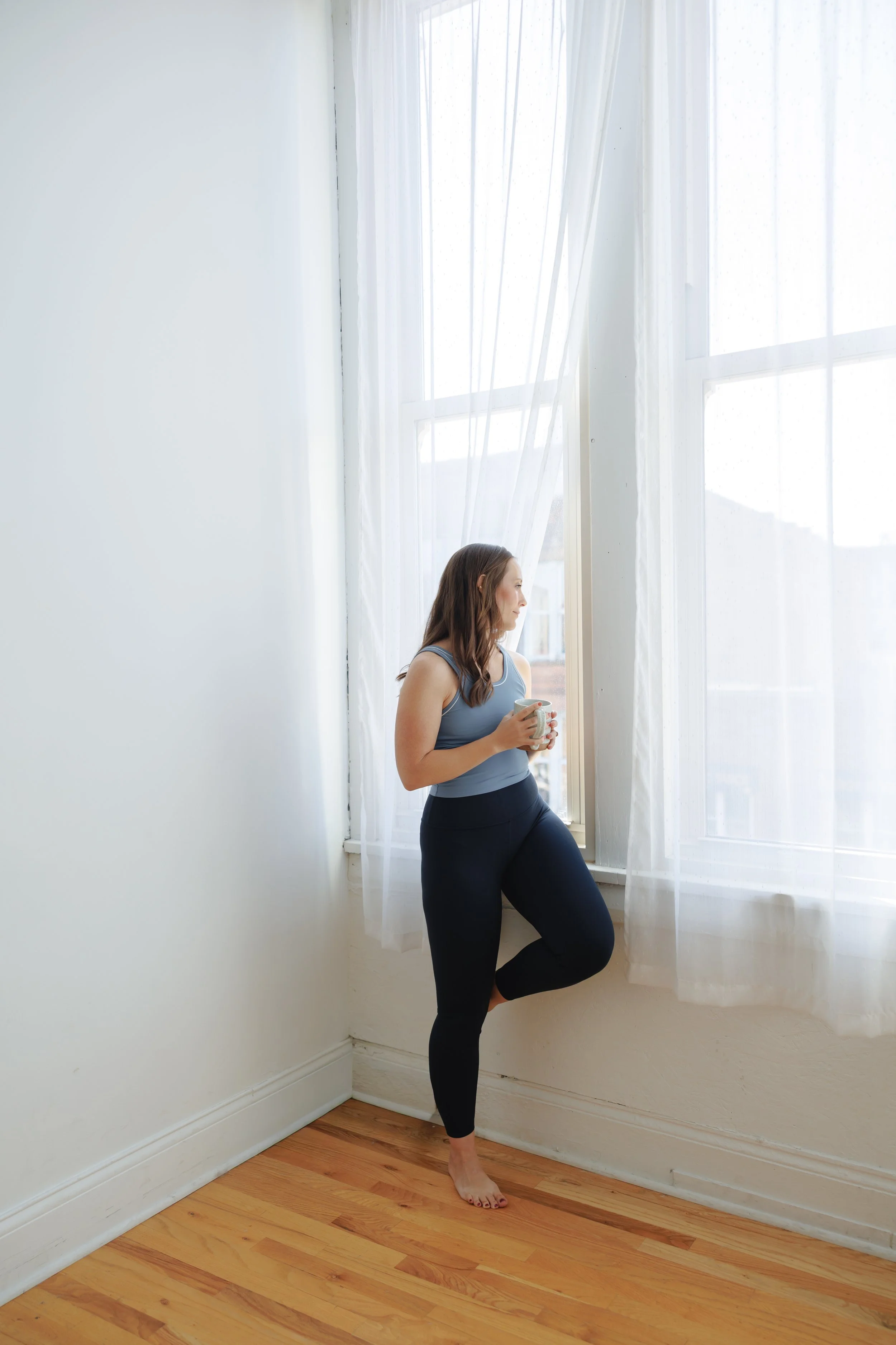 A woman standing barefoot on a wooden floor, looking out a large window with sheer curtains, holding a mug, in a bright room with white walls.