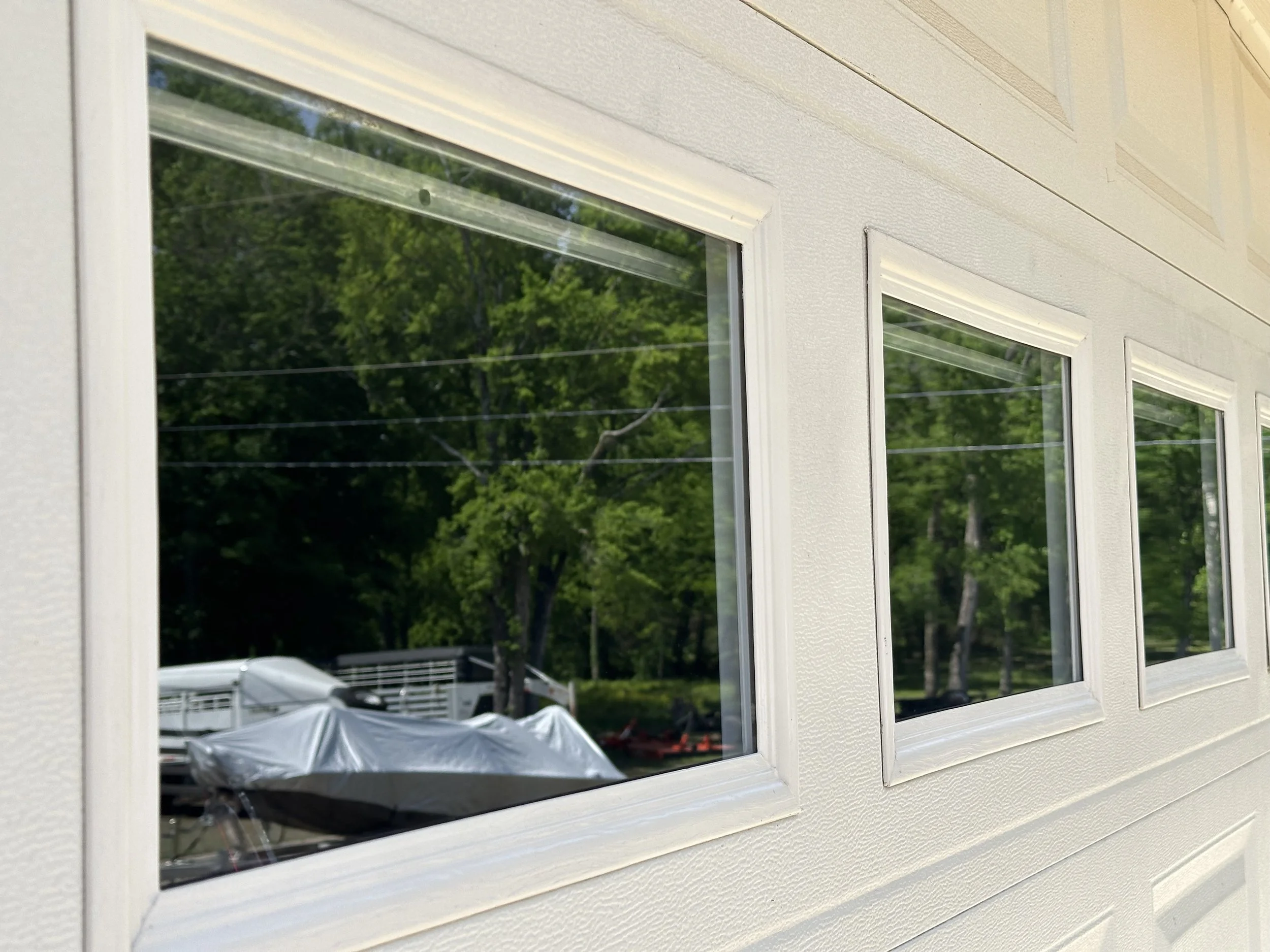Close-up of three white-framed windows on a house exterior, reflecting green trees and a boat covered with a tarp in the yard.