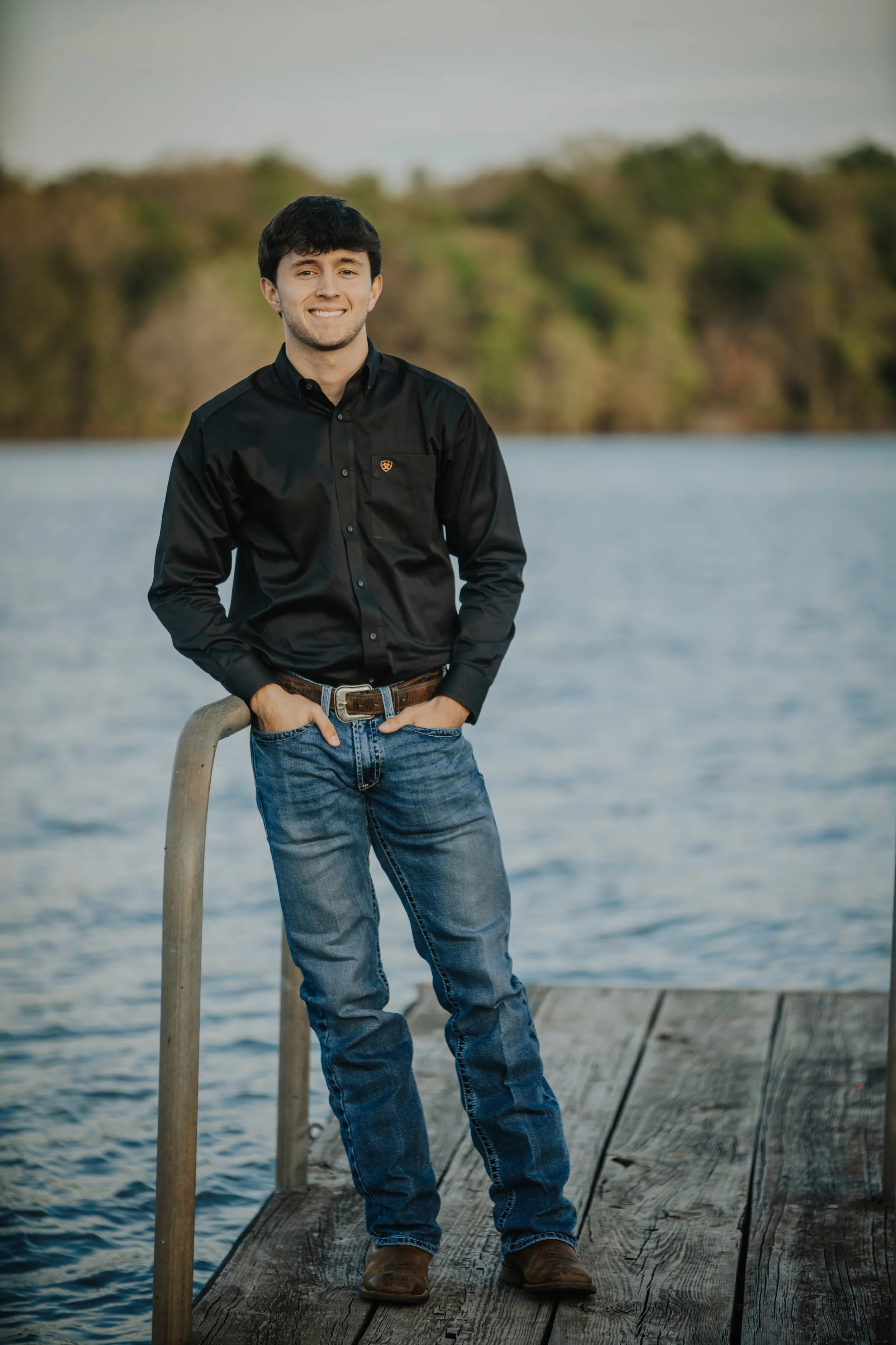 Young man standing on a wooden dock by the water, smiling, with a railing to his side and trees in the background.