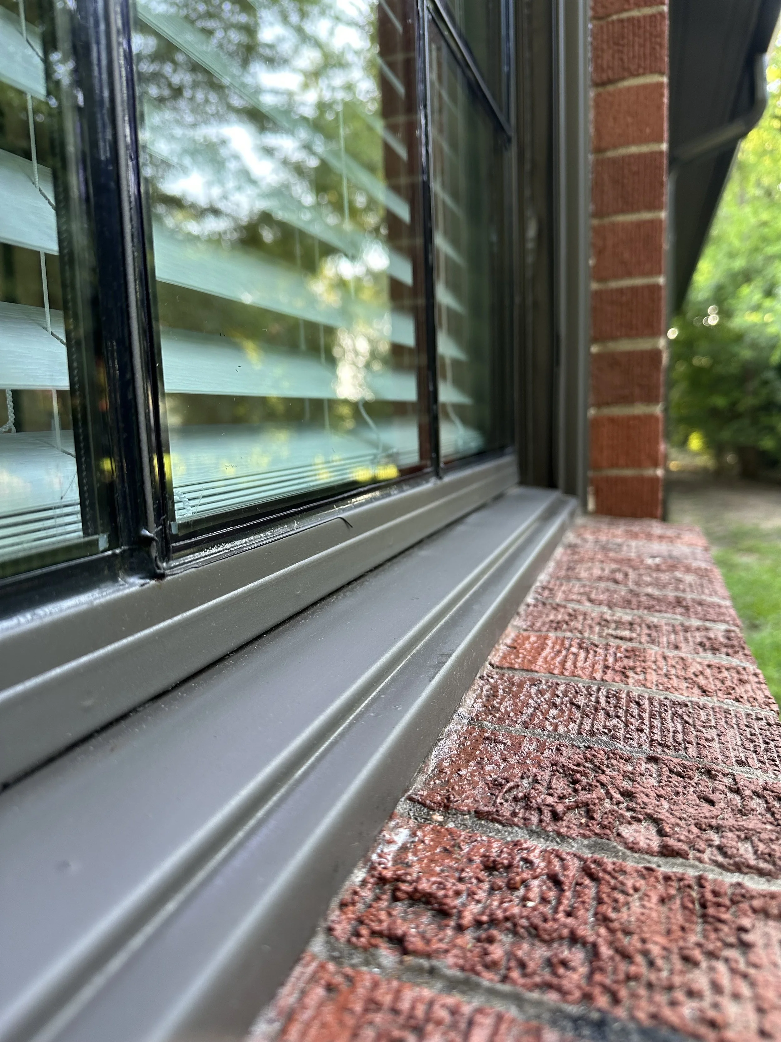 Close-up of a window with blinds, set in a brick wall, with a window sill and greenery reflected in the glass.