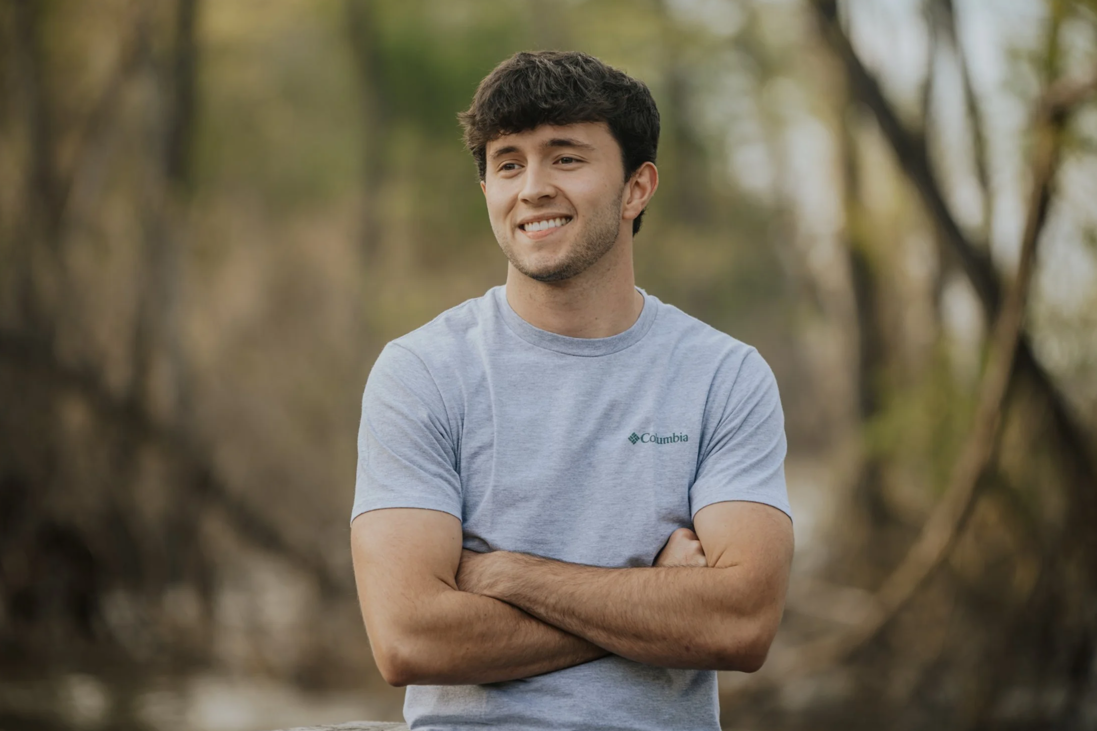A young man with dark curly hair, wearing a gray Columbia t-shirt, smiling with arms crossed, standing outdoors in a forested area.