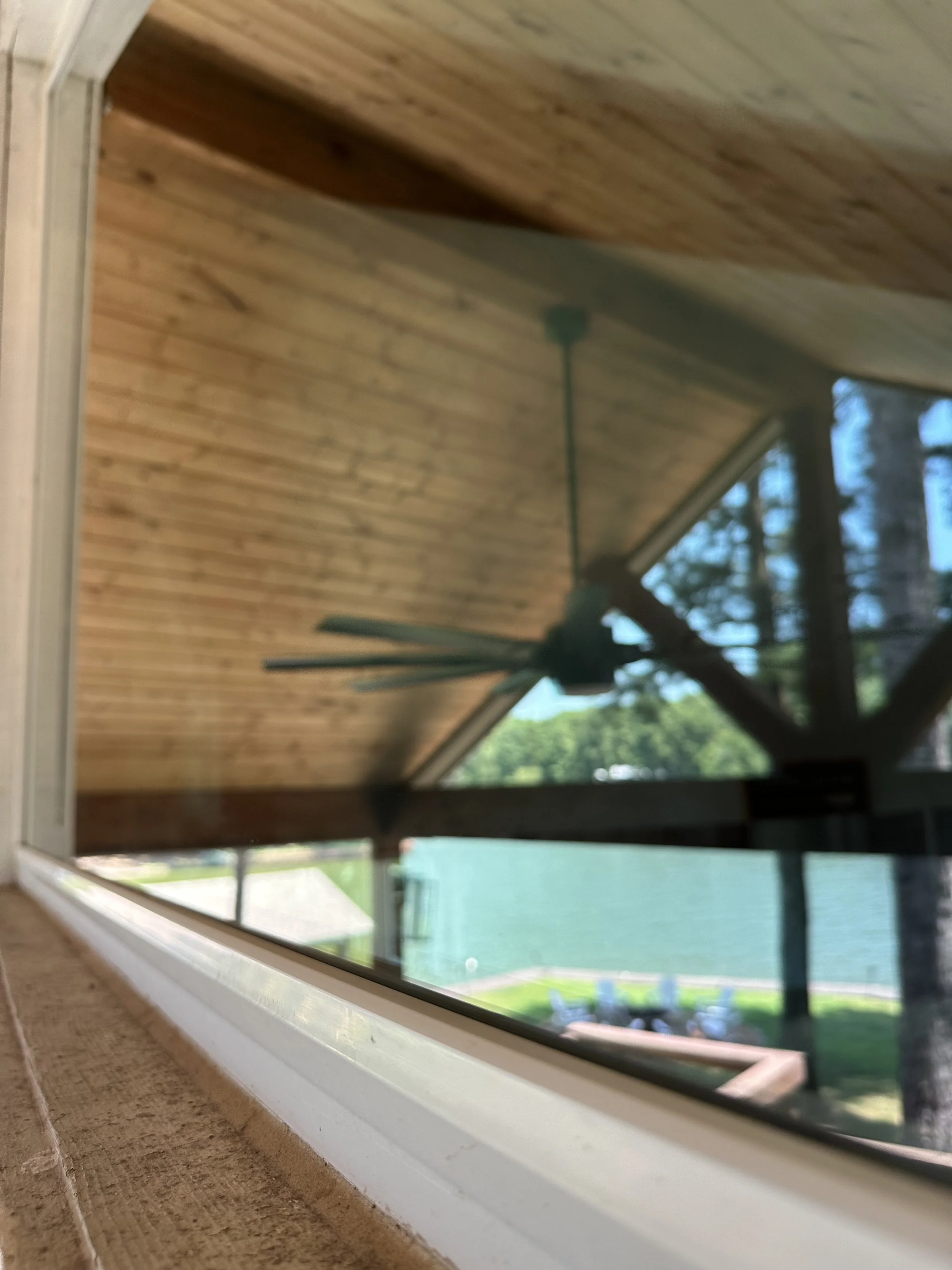 View through a window showing a ceiling fan on a wooden porch roof, trees, and a green shed outside.