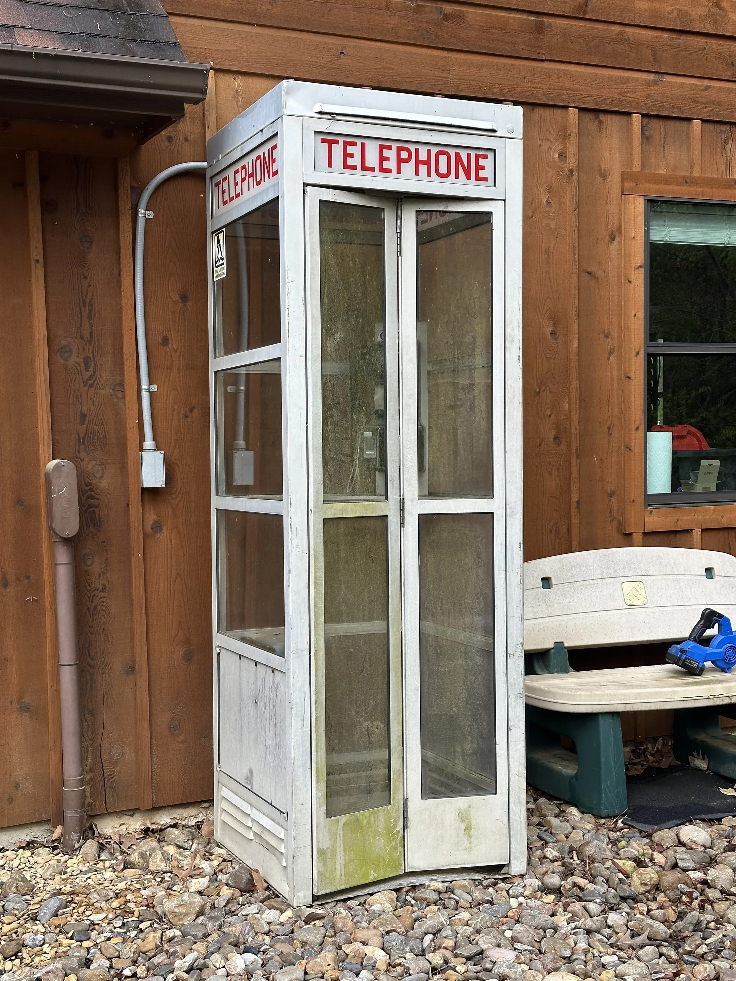 Old outdoor telephone booth with glass panels, labeled 'TELEPHONE' in red, placed on gravel ground beside a wooden building.