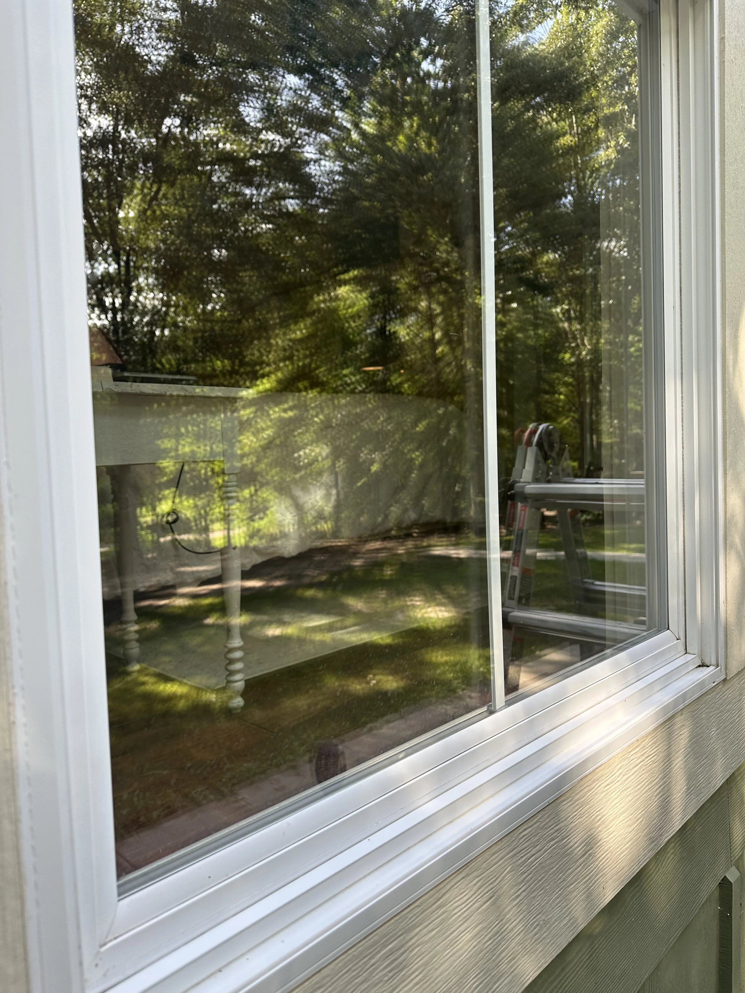 A view through a house window showing trees and greenery outside, with a ladder and some patio furniture visible inside.