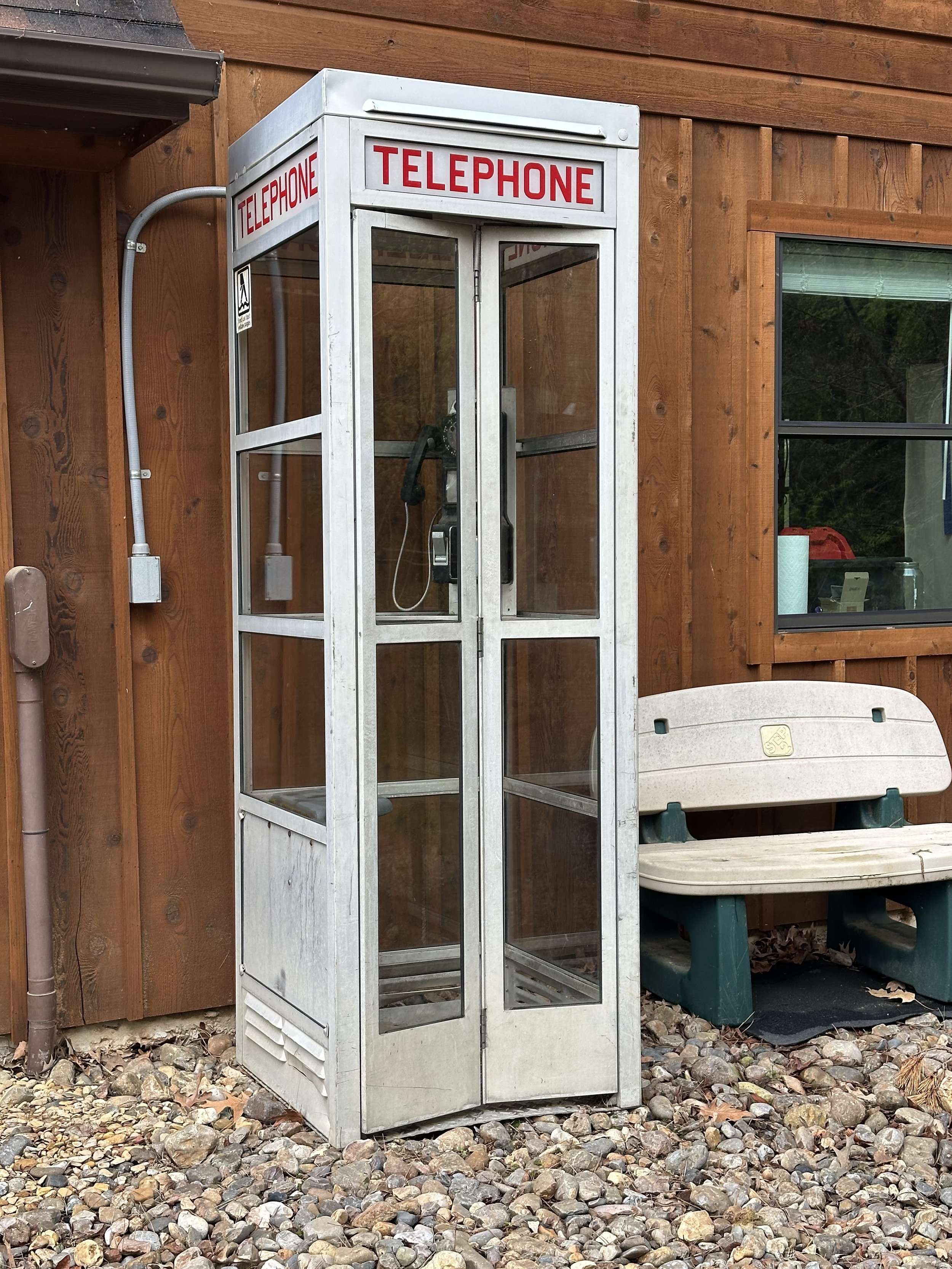 An old metal telephone booth with glass panels, labeled 'TELEPHONE' in red, placed outside on a gravel surface next to a wooden building. A bench is nearby.