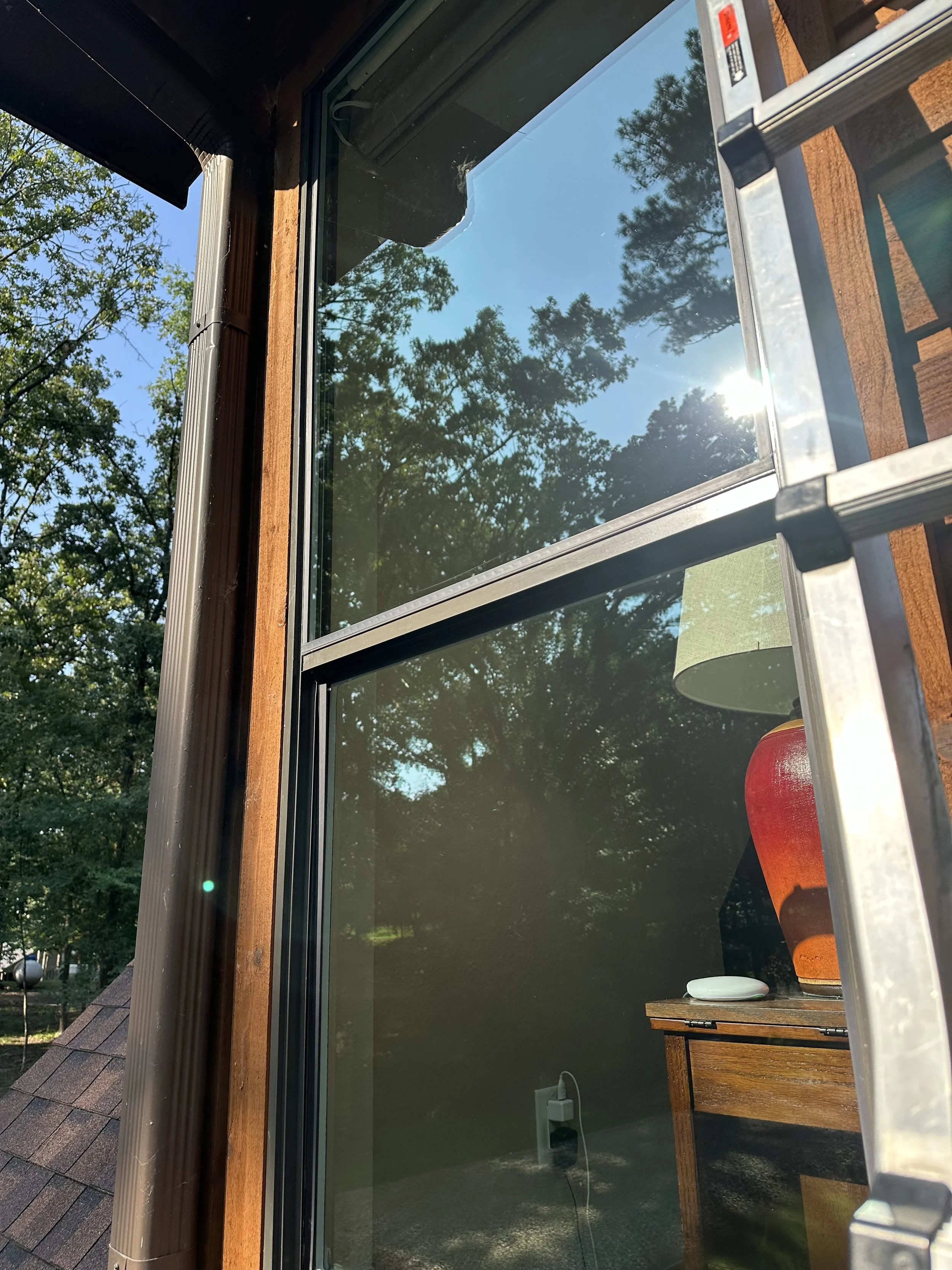 Close-up of a window reflecting blue sky and trees outside, with interior showing a red-orange lamp and a wooden table.
