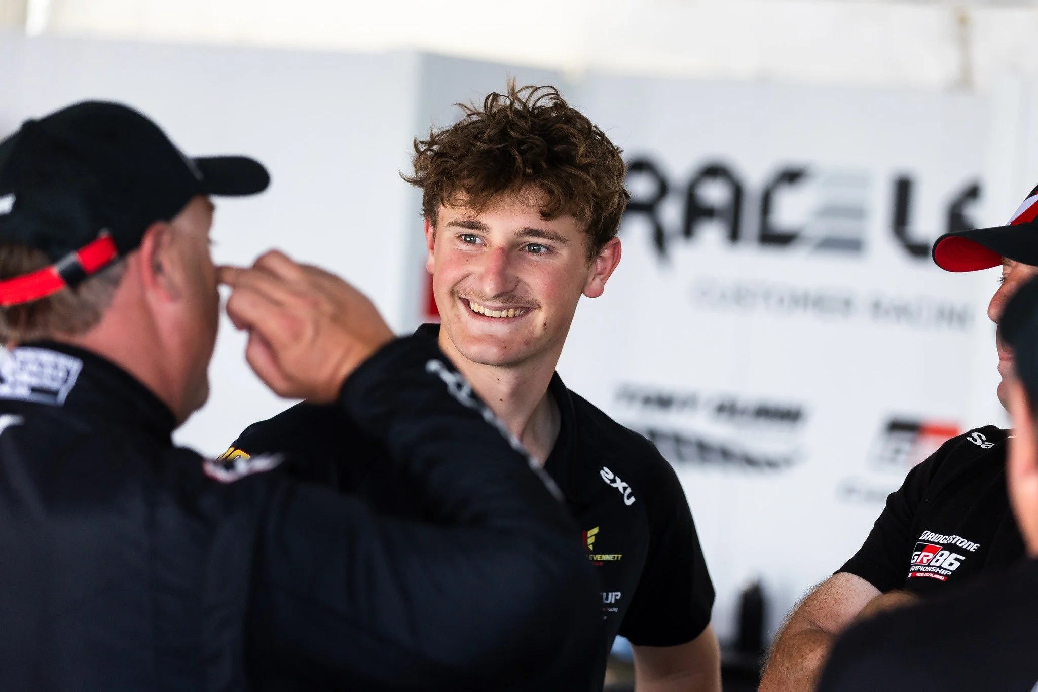 Lachlan Evennett is seen smiling, engaged in conversation with three other people, in what appears to be a racing team garage or pit area.