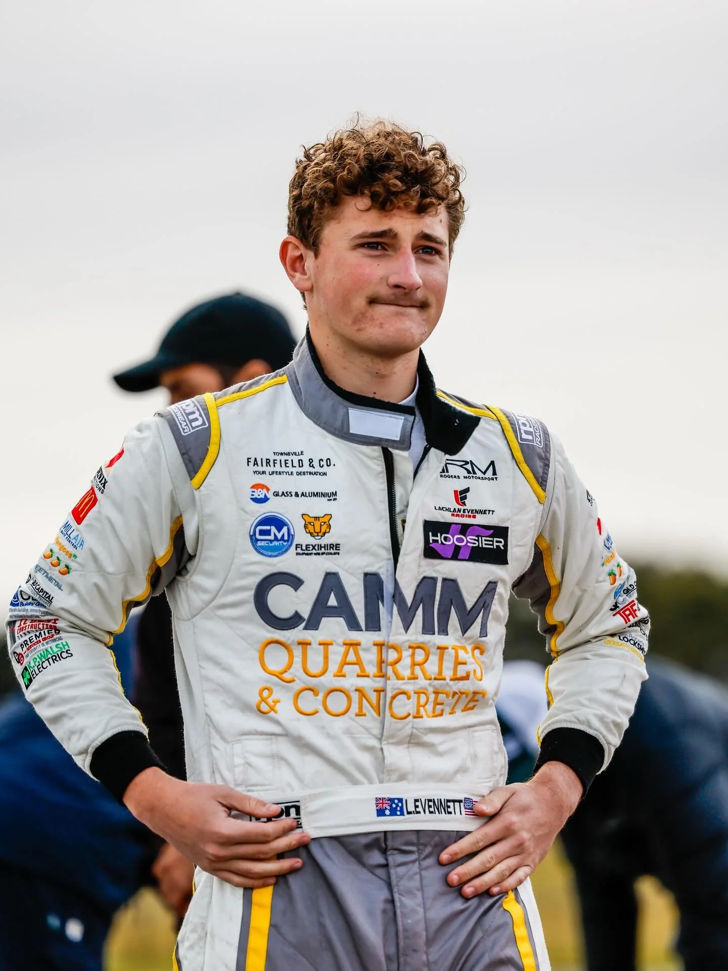 A picture of race car driver Lachlan evennett in a white racing suit with various sponsor logos, standing outdoors with a cloudy sky in the background. He has curly hair and a contemplative expression.