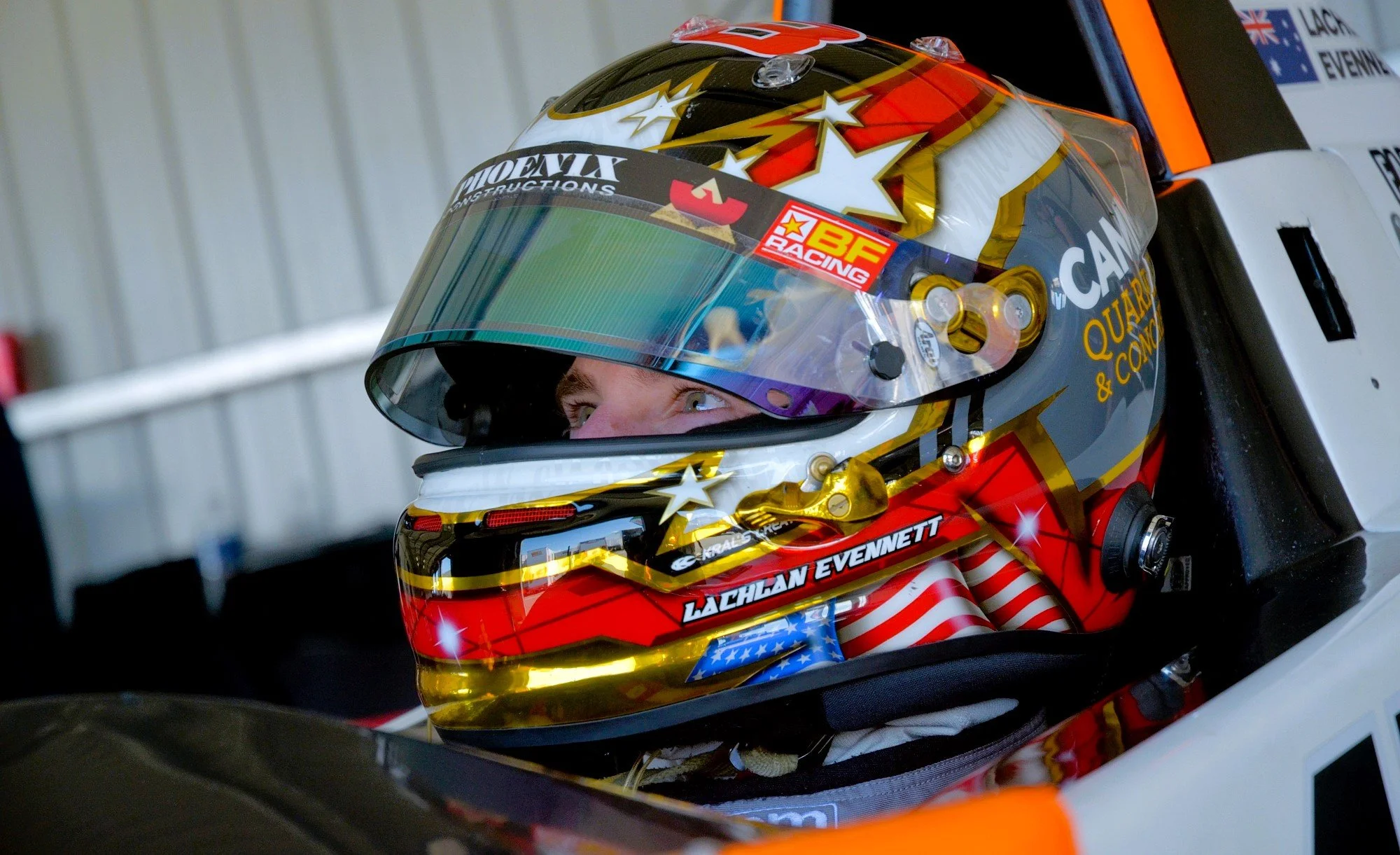 Close-up of a race car driver wearing a decorated helmet with stars, stripes, and gold accents, sitting in a racing car.