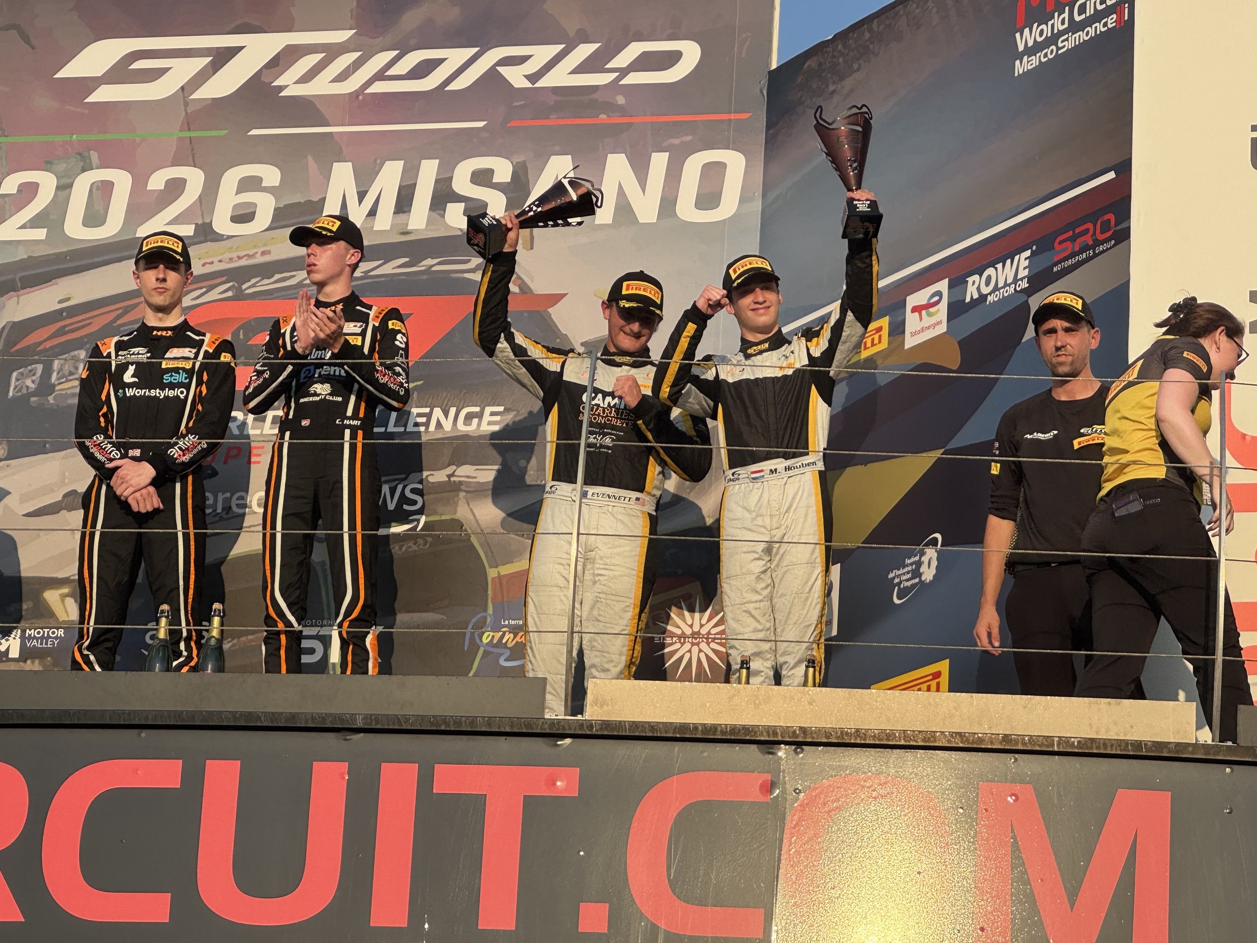 A group of race car drivers on a podium celebrating their victory, holding trophies, with sponsor logos and a large banner in the background.