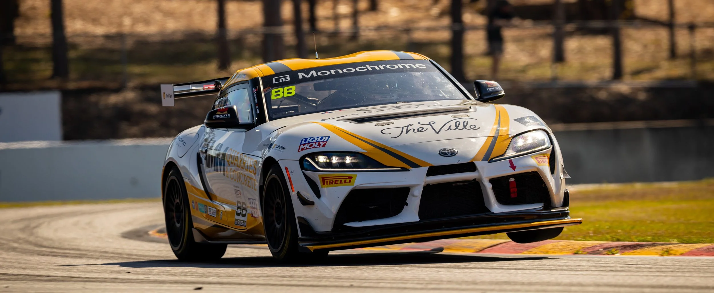 A white and yellow race car with the number 88 on the windshield, speeding on a race track, surrounded by a blurred background of trees and fencing.