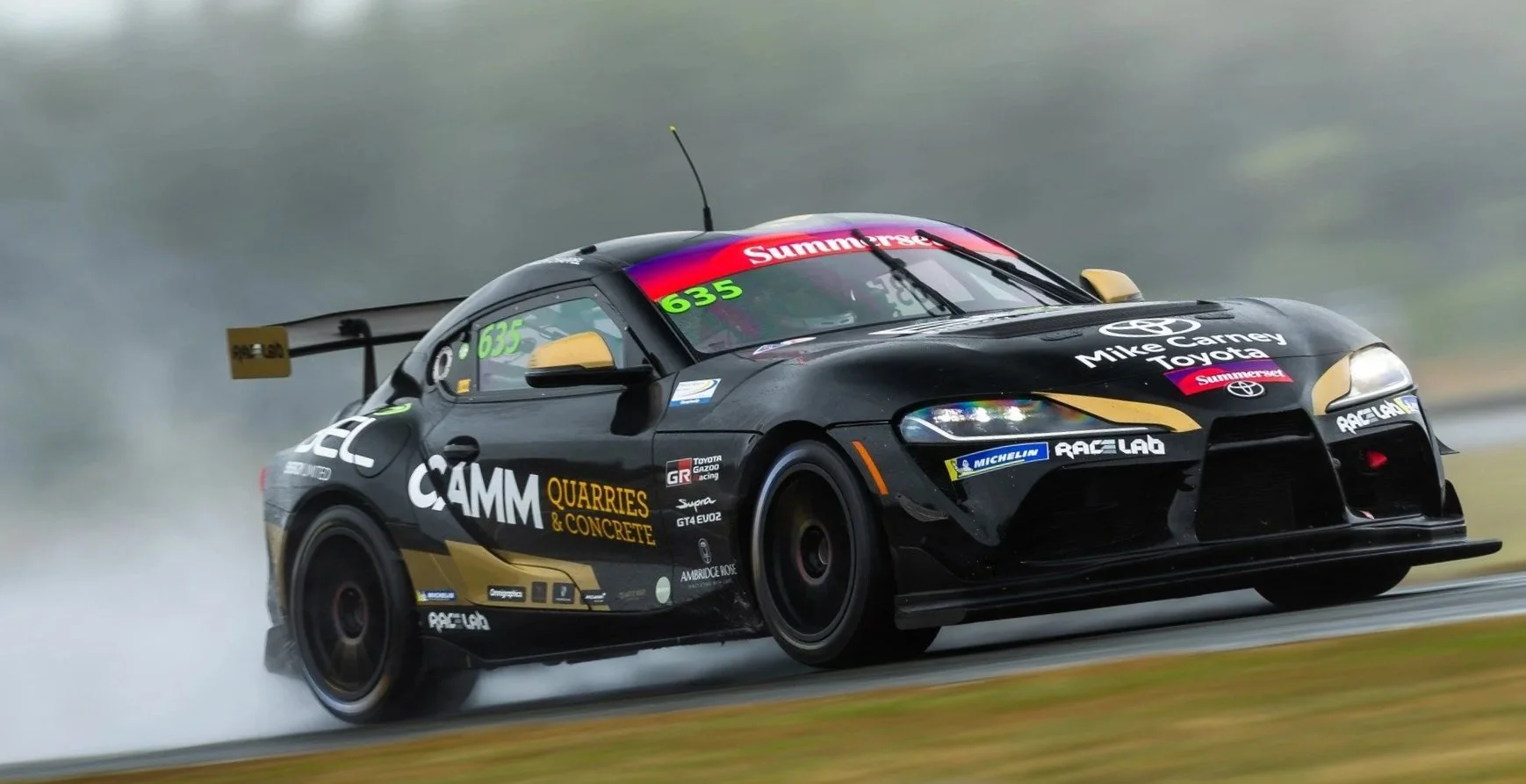 A black race car with yellow accents speeding on a wet track, creating a spray of water behind it, with various sponsor logos and the name Mike Carney on the hood.