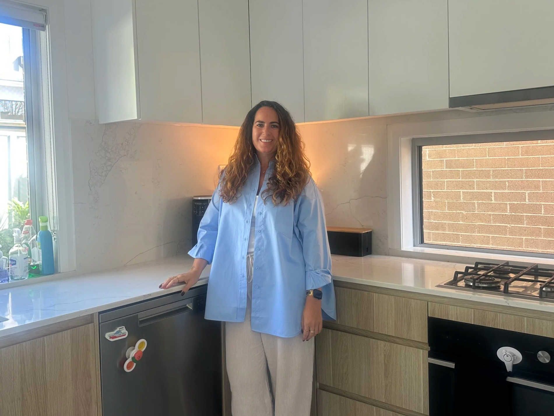 Laura, A Simplified State founder, stands in a modern kitchen near a window, wearing a light blue shirt and beige pants, smiling at the camera.