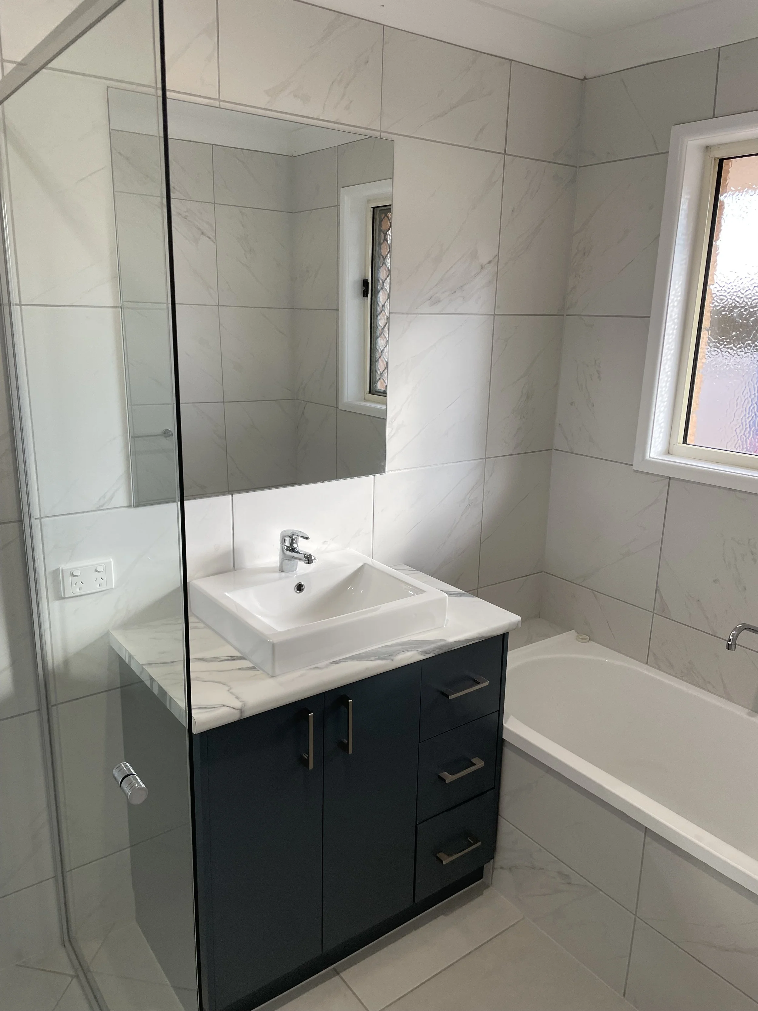 Modern bathroom with white marble tiles, a mirror above a white rectangular sink, a black vanity cabinet, a bathtub, and frosted windows.