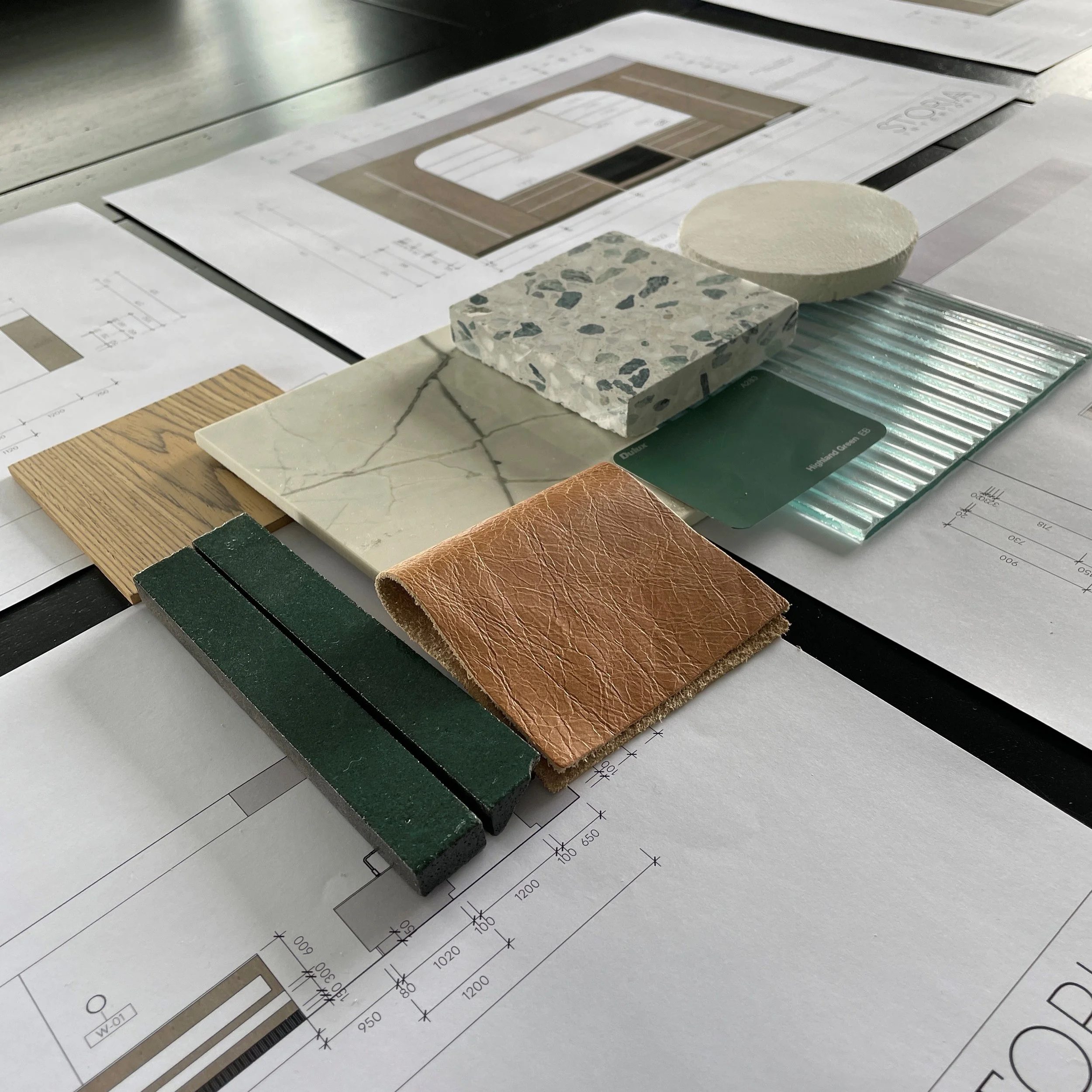 Interior design material selection for a Melbourne project, showing a palette of tiles, wood, and leather samples on architectural blueprints by Storia Interiors.