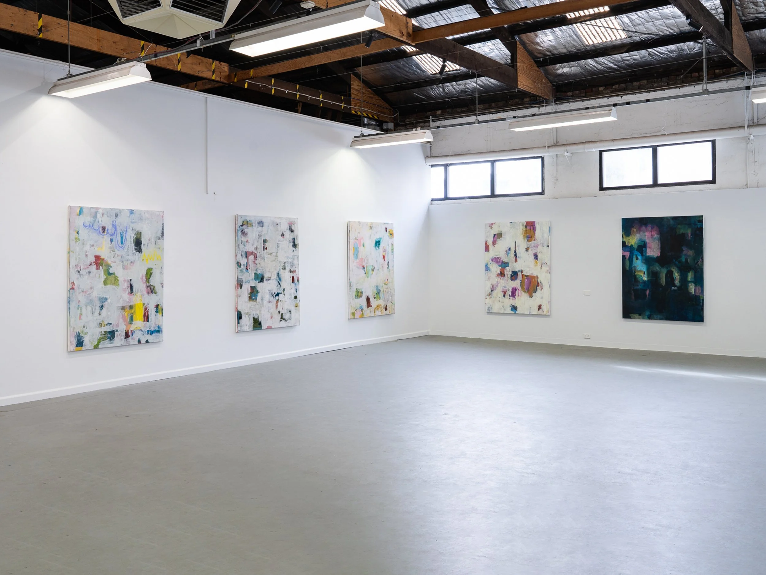 An art gallery with five colorful abstract paintings hung on a white wall. The gallery has a high ceiling with exposed beams and windows near the ceiling letting in natural light.
