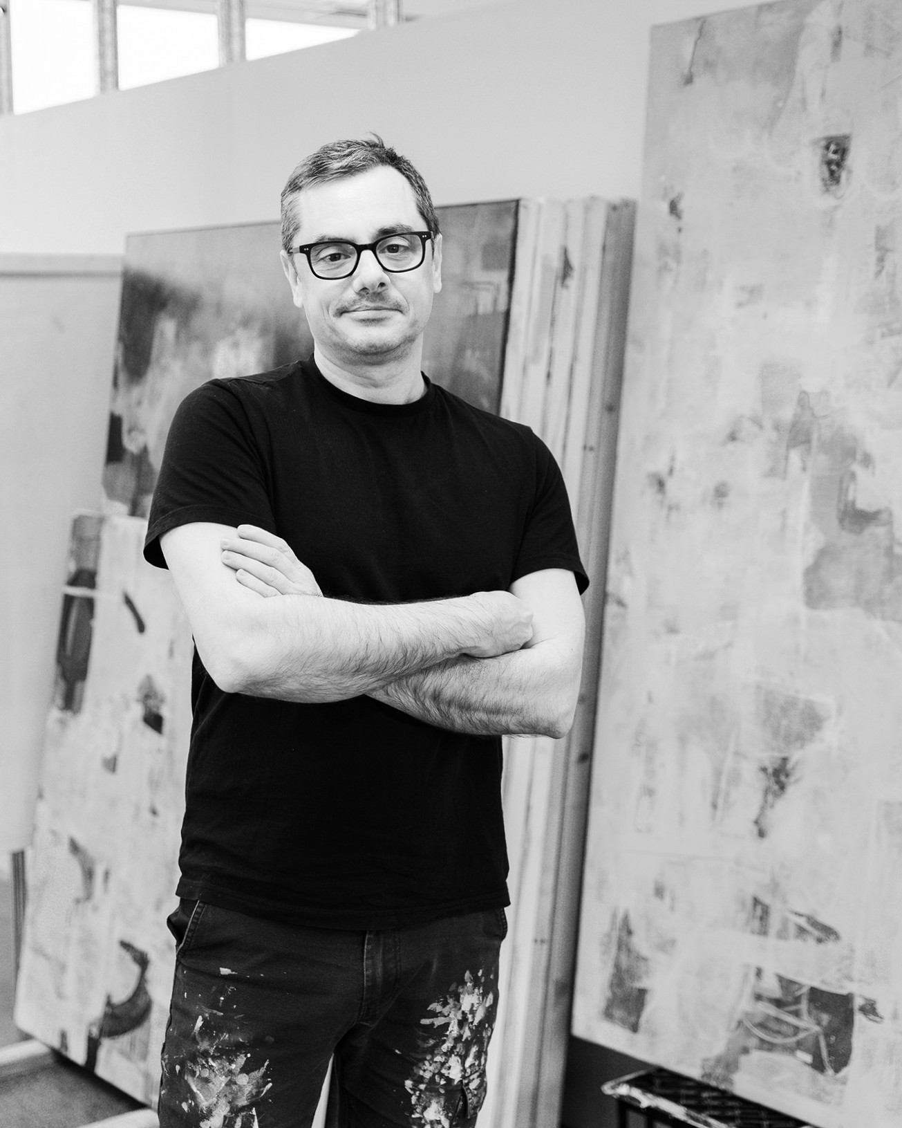 A man wearing glasses, a black t-shirt, and paint-splattered pants standing with arms crossed in an art studio, with canvases and painting supplies in the background.