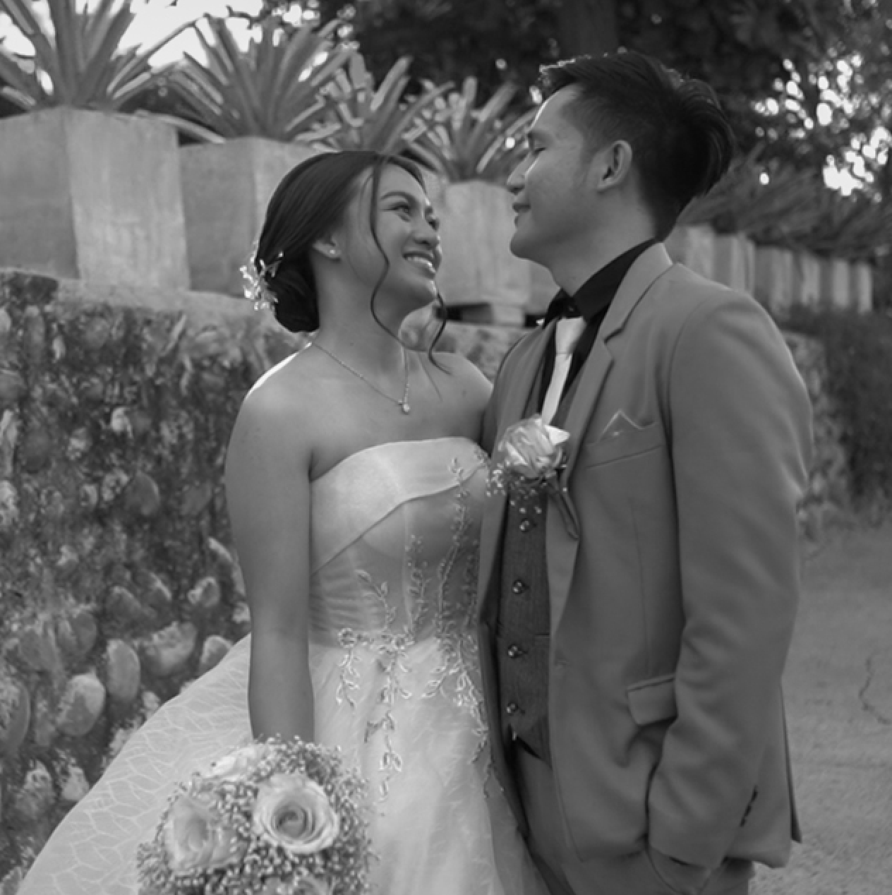 A couple dressed in wedding attire gazing into each other's eyes outdoors, with plants and stone steps in the background.