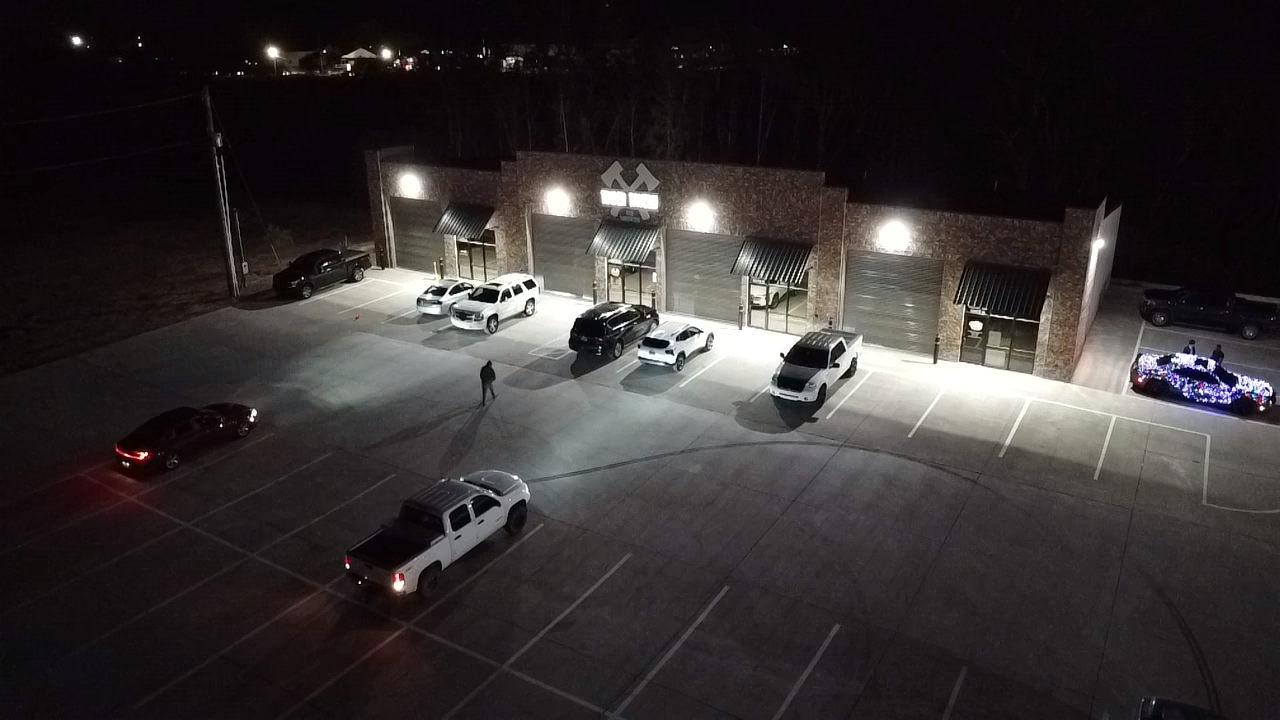 Nighttime view of a building with multiple garage doors and a sign above, a few cars parked in front, one person walking across the parking lot, and a decorated vehicle with lights on the far right.