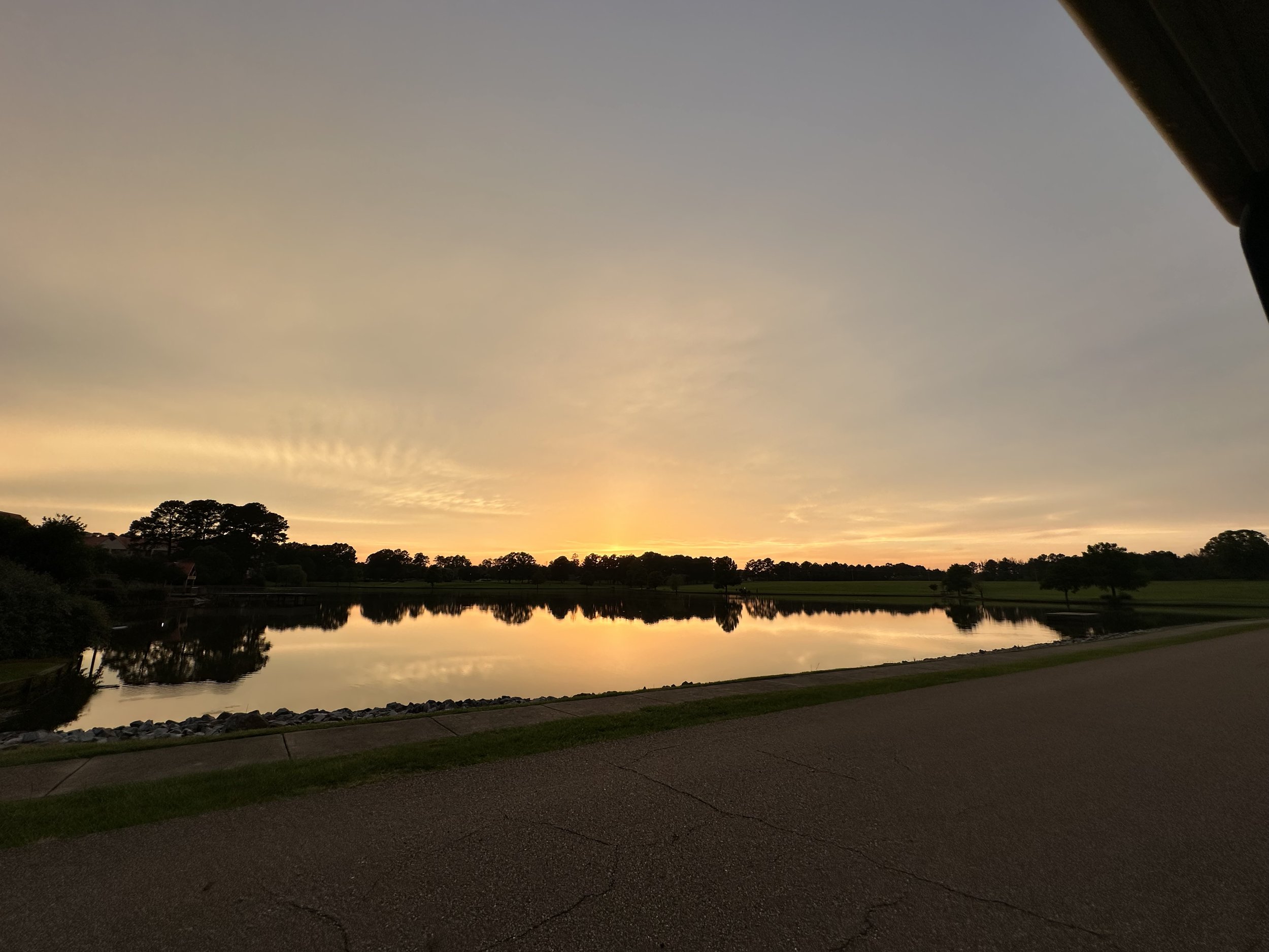 Sunset over a calm lake with trees and green fields reflected on the water, a paved pathway in the foreground.
