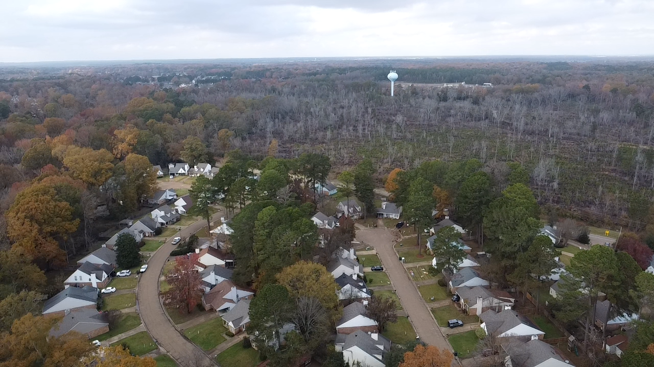 An aerial view of a residential neighborhood with houses, trees, and roads, with a water tower in the background and a forested area.