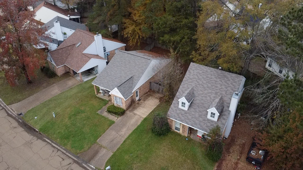 Aerial view of three houses with driveways and surrounding trees, some with fall foliage, in a suburban neighborhood.