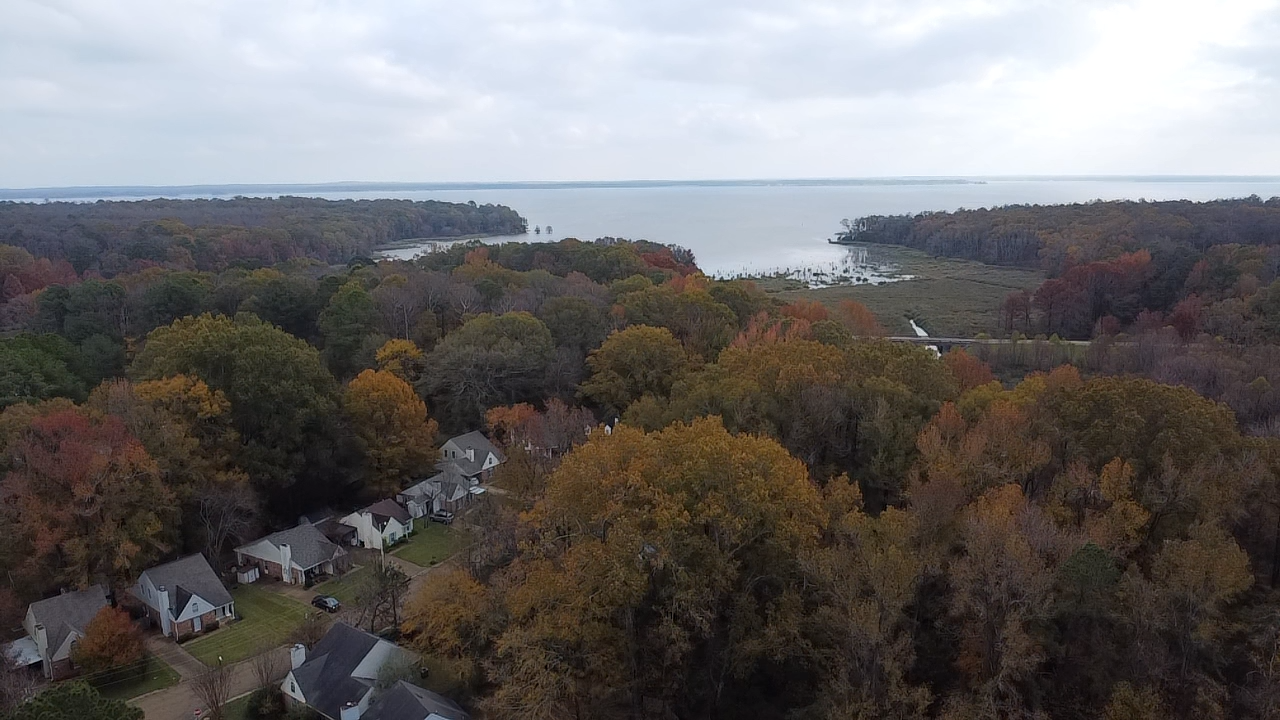Aerial view of a residential neighborhood with houses surrounded by fall-colored trees, overlooking a lake with a tree-lined shoreline and a cloudy sky.