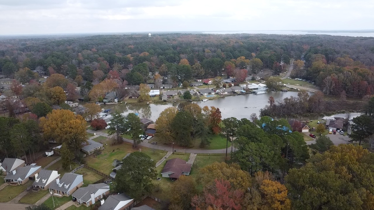 Aerial view of a neighborhood with a lake, houses, trees in fall colors, and a wooded area in the background.