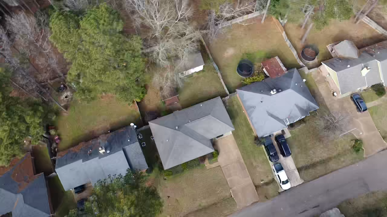 Aerial view of a suburban neighborhood showing several houses with driveways, lawns, and trees in the backyard, some with backyard structures and fences.