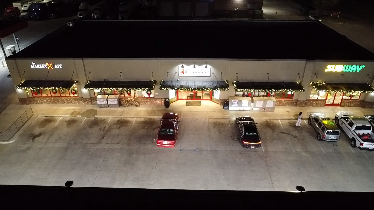 A strip mall at night with parking lot and four parked cars, featuring a Market X Cafe and Subway restaurant, decorated with holiday lights and wreaths.