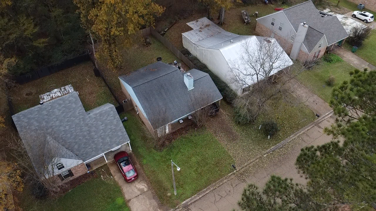Aerial view of a residential neighborhood showing three houses with lawns, trees, cars, and a sidewalk, during fall.
