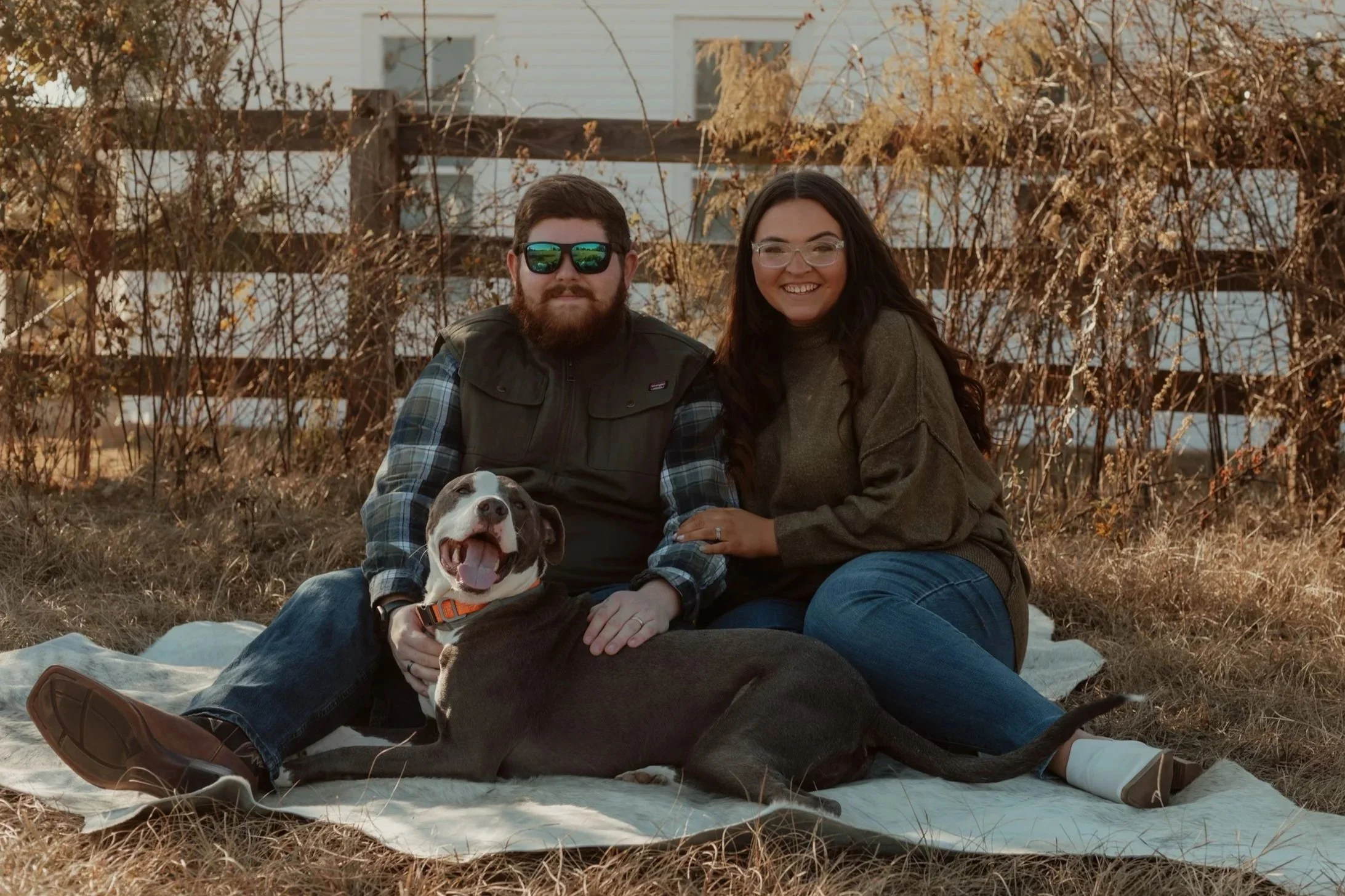 A smiling couple sitting on a blanket outdoors with their dog, in front of a wooden fence with dried foliage, during autumn.