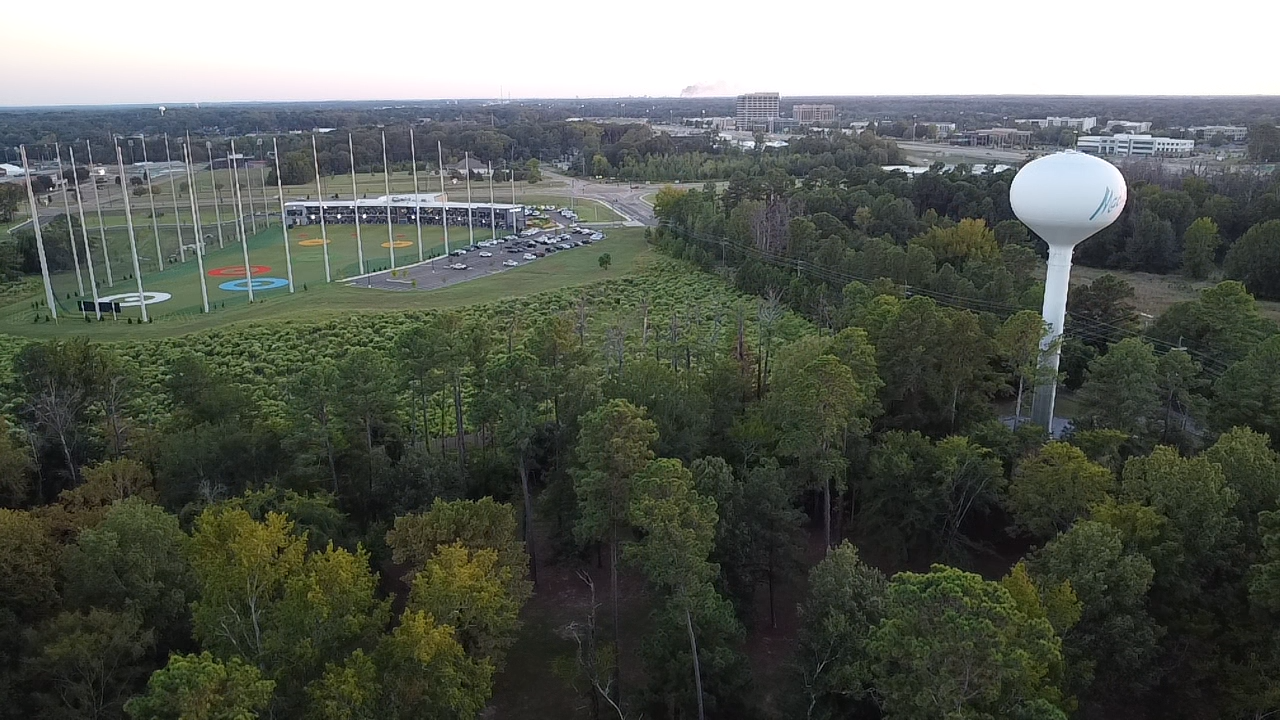 Aerial view of a green park with a baseball field, parking lot, and a large white water tower in a wooded area with trees, followed by city buildings in the distance.