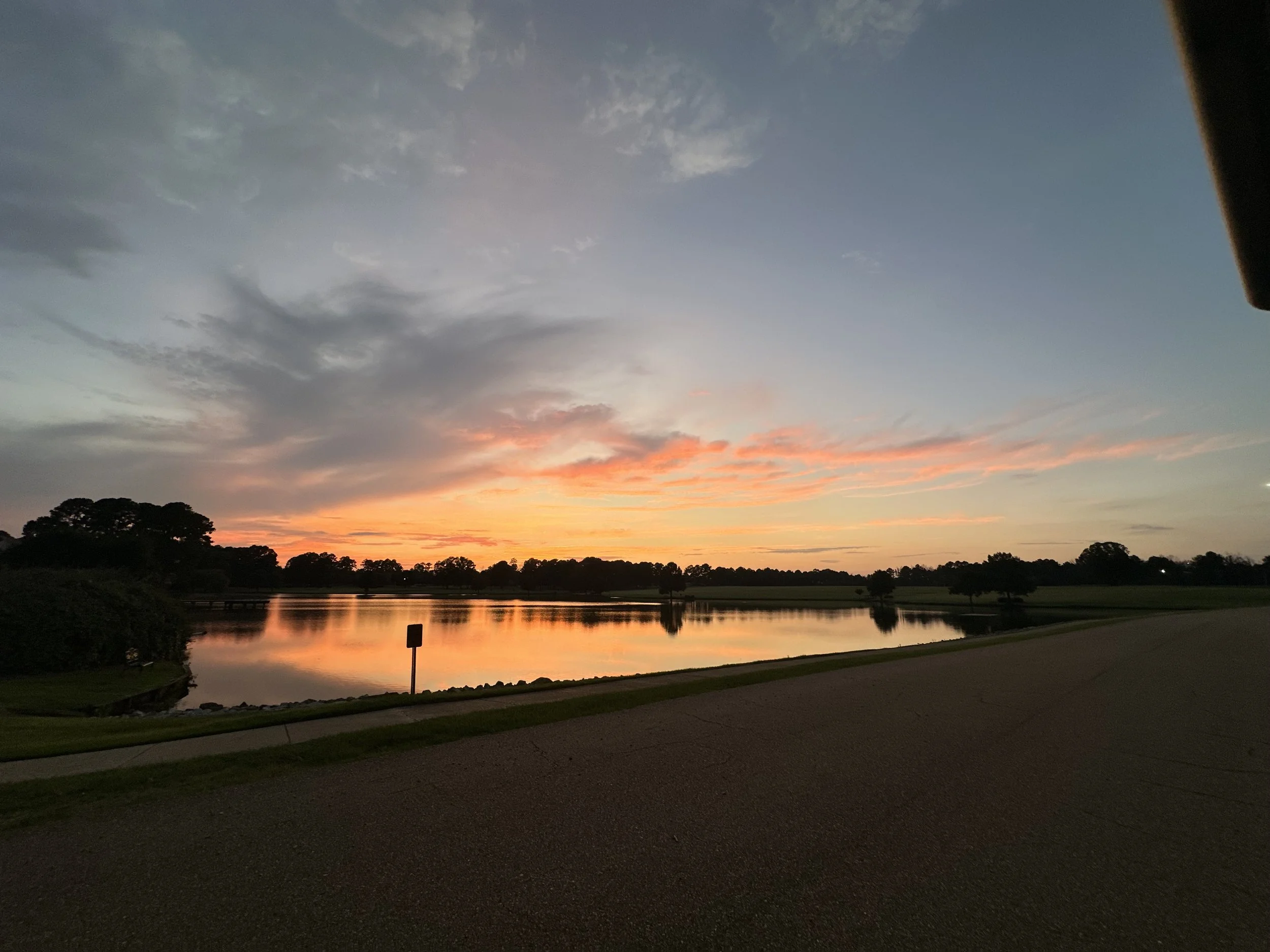 Sunset over a lake with a partly cloudy sky, silhouetted trees, and a sidewalk in the foreground.