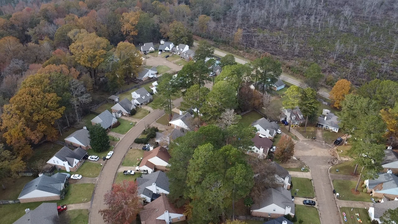 Aerial view of a suburban neighborhood surrounded by trees with fall foliage, featuring single-family homes, driveways, and cars parked on the streets and in yards.