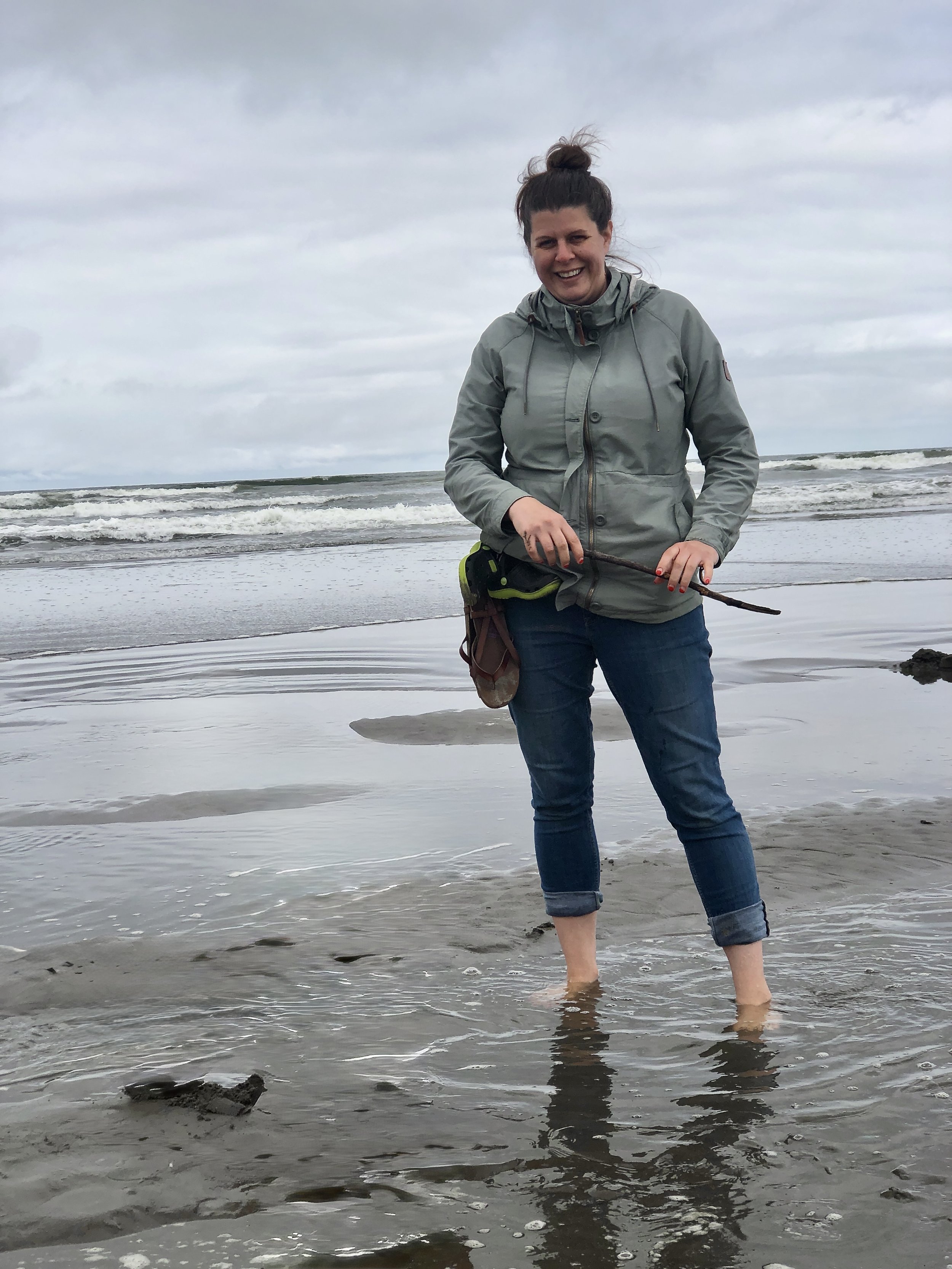Monica Vasquez standing on the beach at the Oregon Coast, smiling and holding a stick near the ocean.