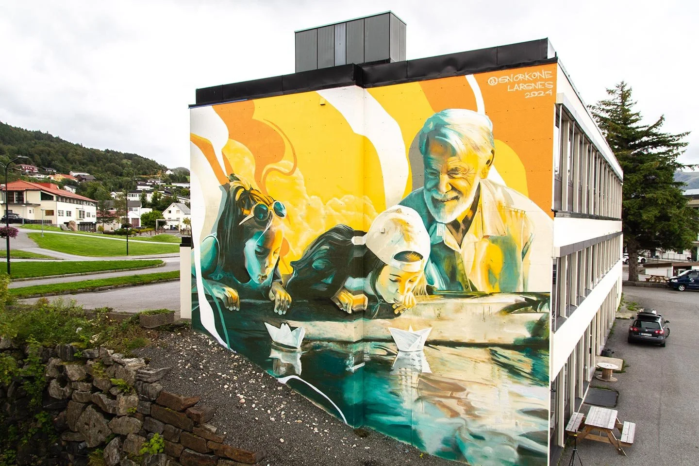 Gatuväg med ett stort väggmålning av en glad man nära vatten, barn och hund, med flytande papperbåtar i elva, i en stadsmiljö.