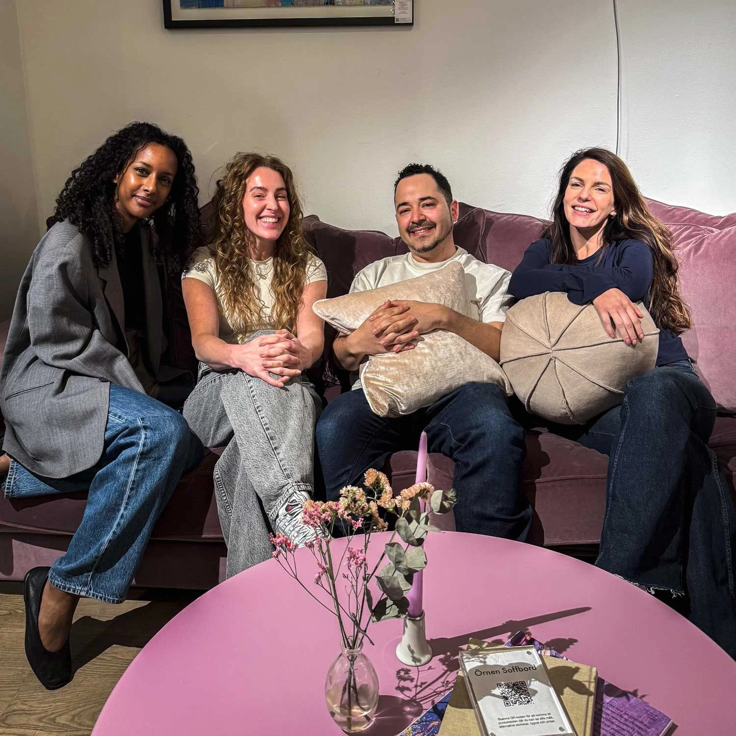 Fyra personer sitter på en soffa och skrattar, tre kvinnor och en man, i ett mysigt inomhusrum med ett bord med blommor framför.