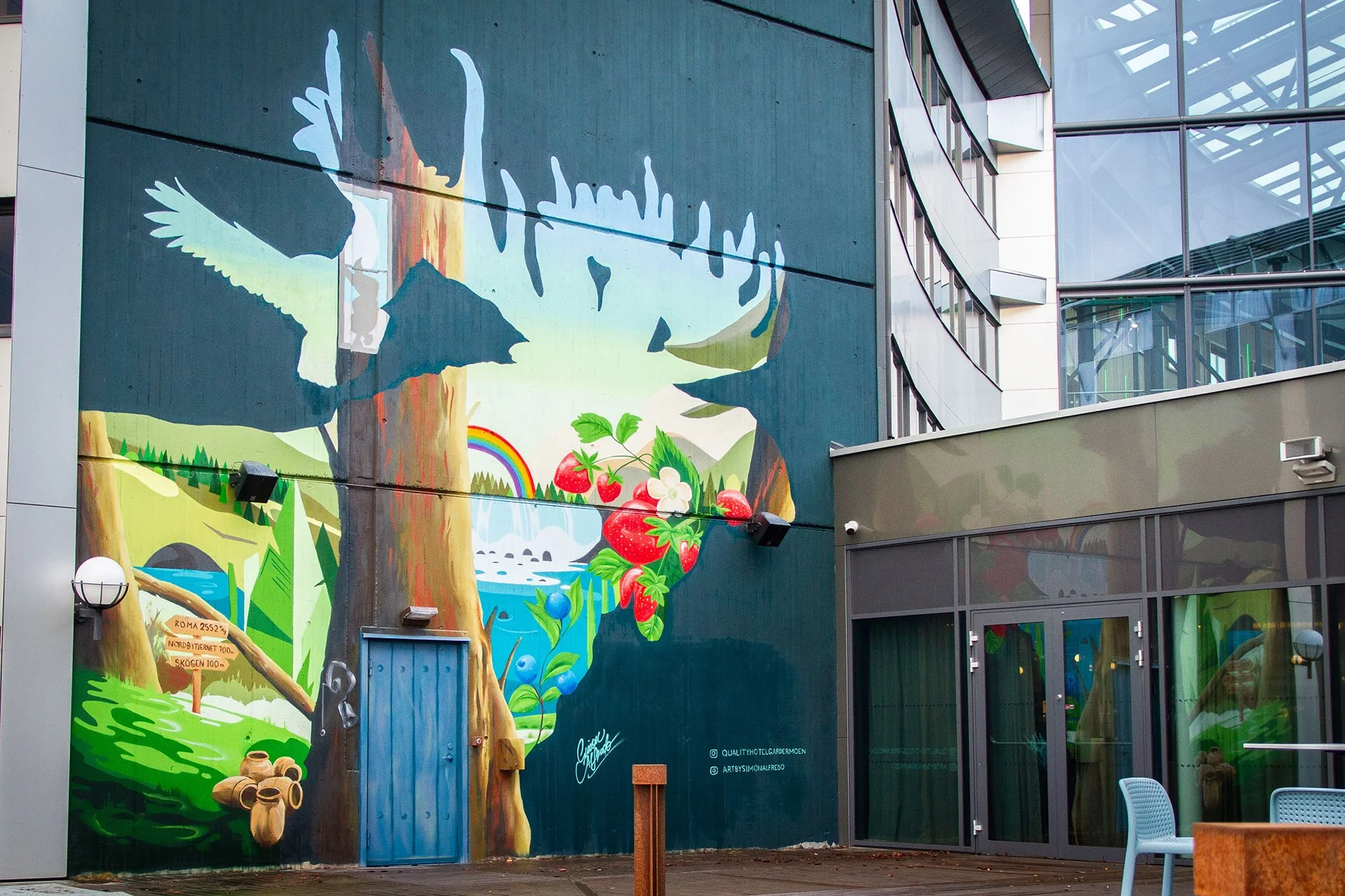 Färgglad väggmålning av ett skogsmiljö med träd, en bäck, regnbåge och stora jordgubbar.