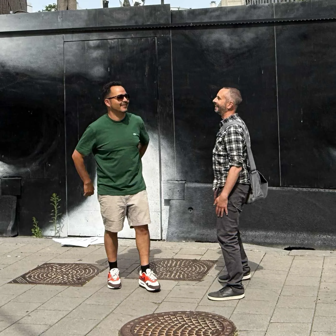 Två män pratar med varandra på gatan, en i grönt t-shirt och shorts, den andra i rutig skjorta och byxor, med bakgrund av en svart vägg.