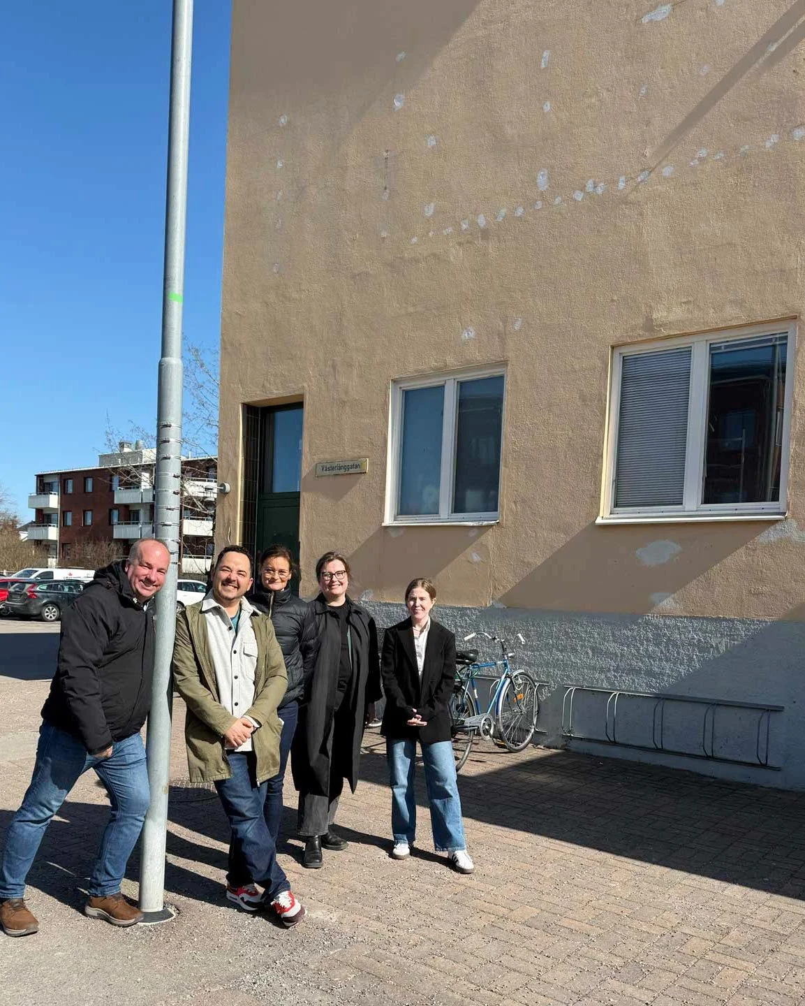 Fem personer står bredvid ett gatuhörn med en vägg med några fönster, ett cykelställ och en gatlampa, i ett bostadsområde under en klar himmel.