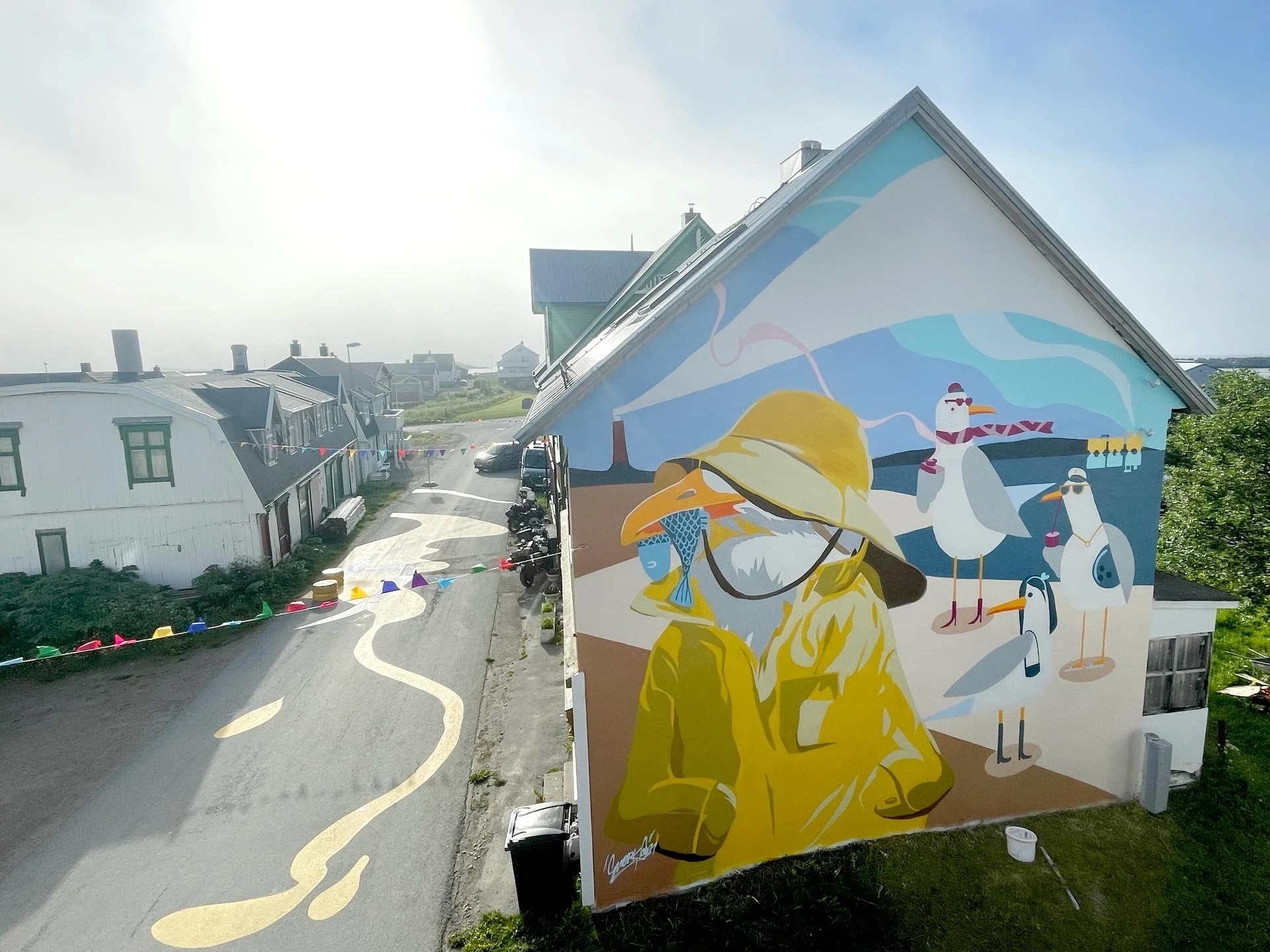 Gatuvy med en stor väggmålning av en person med solhatt och solglasögon, samt några måsar i fågelillustrationer, på en byggnad i en liten stad eller by.