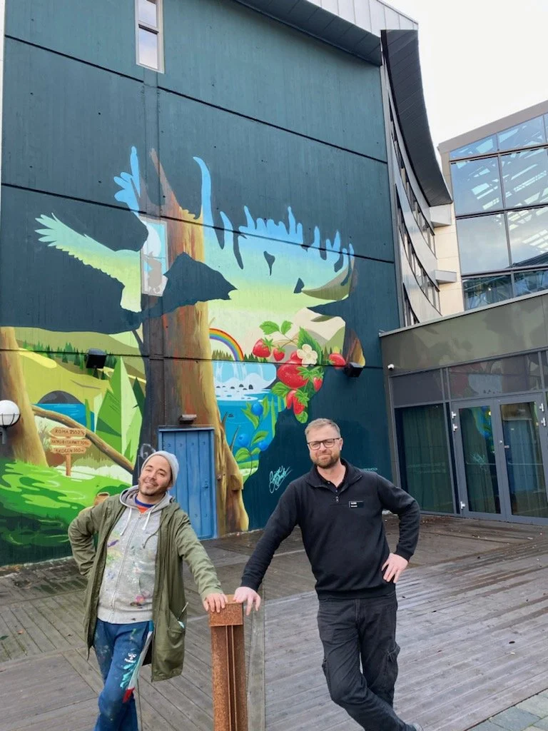 Två män poserar framför en stor muralmålning av ett naturlandskap med träd, en flod, en regnbåge och frukt på en vägg utomhus.