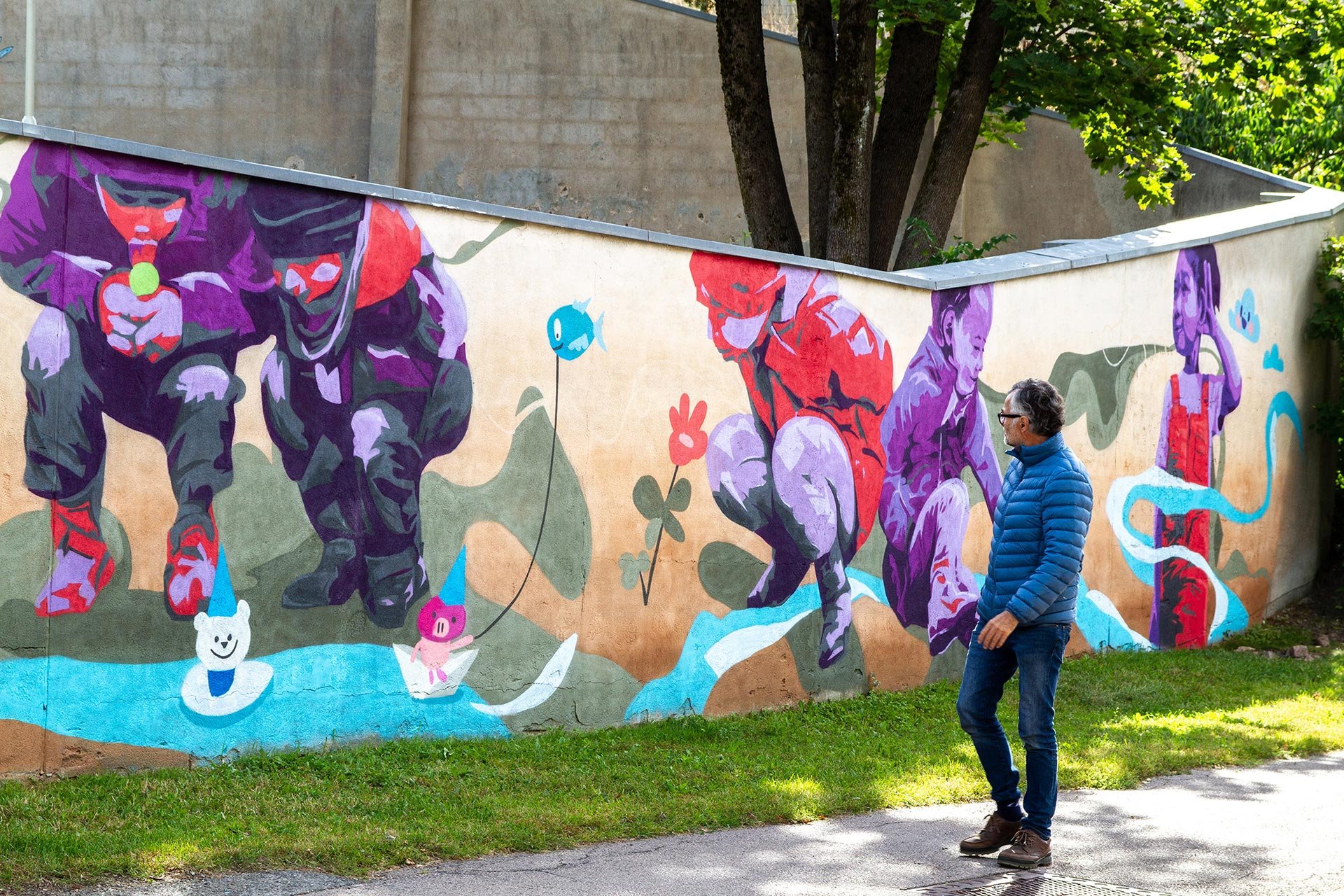 Gatuväggmålning med färgglada djur och människor vid en flod, en man i blå jacka går på trottoaren framför målningen.