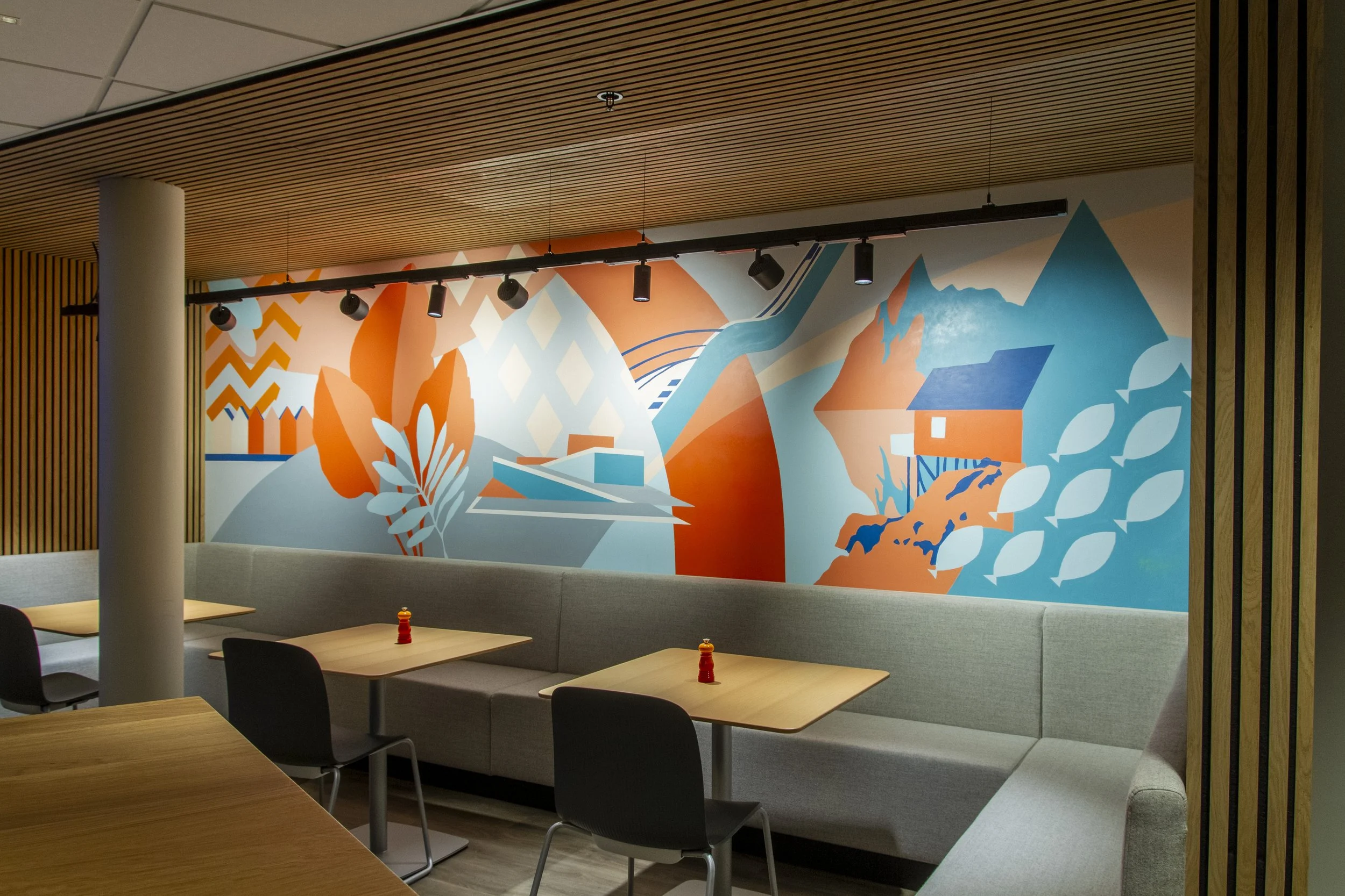 Färgglad väggmålning med landskap, fjäll, vatten och hus i orange, blå och gröna nyanser i ett modernt inredningsrum.