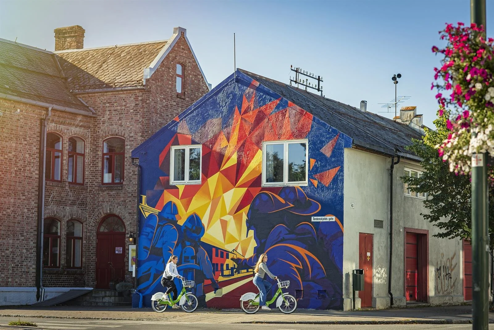 Färggrant gatukonst som avbildar brandmän i blåa uniformer och hjälmar, med brinnande byggnader i bakgrunden, på en vägg på en stadsgata, medan två personer cyklar förbi