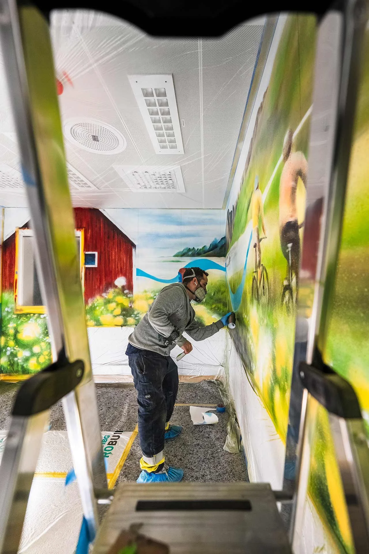 Simon Compagnet Diaz med en muralmålning med en cyklist och ett landskap, han bär skyddsutrustning och använder sprayfärg,